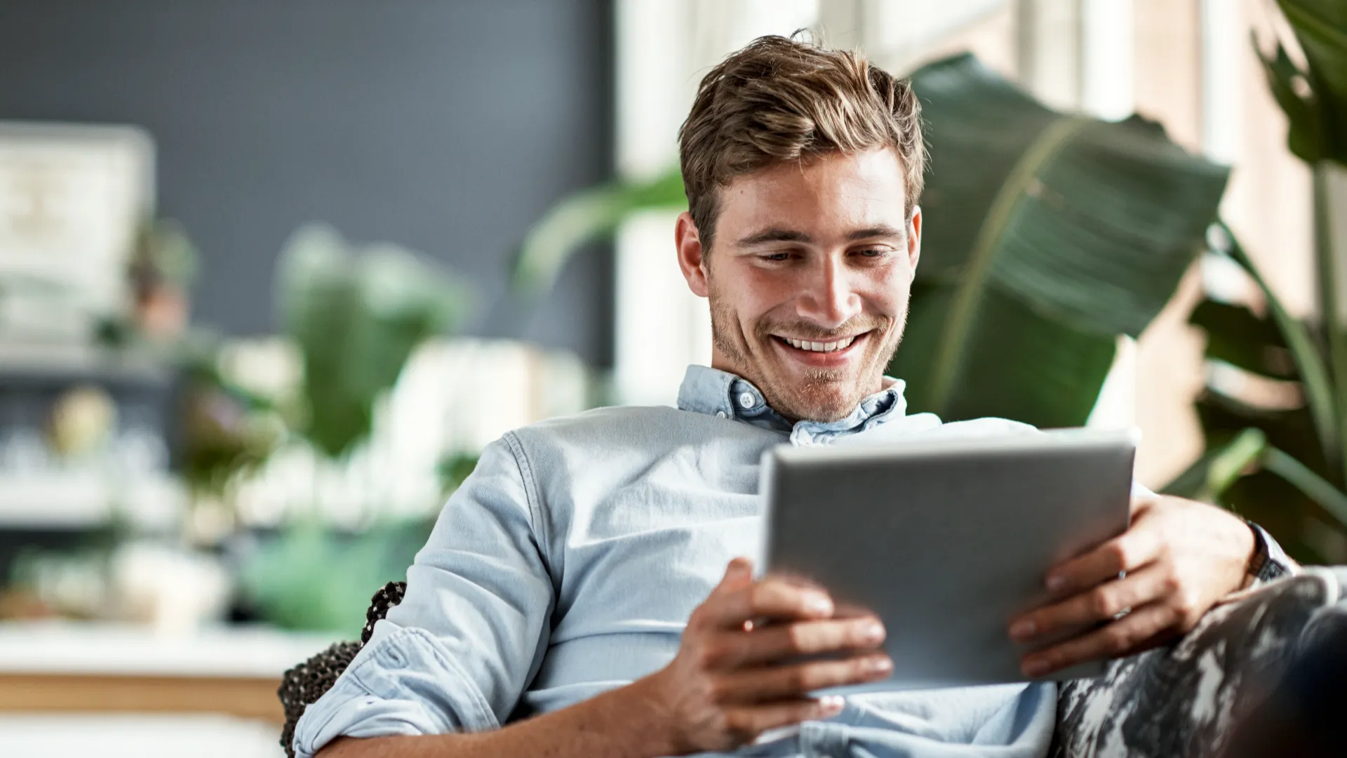 Person sitting on a couch, using a tablet in a relaxed indoor setting with greenery in the background, illustrating casual technology use