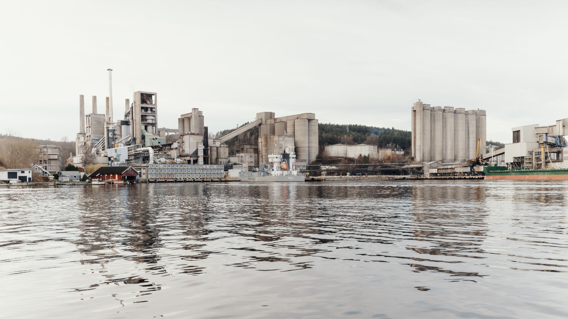 A waterside industrial facility featuring large silos, chimneys, and a docked ship, surrounded by trees and hills – part of Heidelberg Materials’ carbon capture initiative.