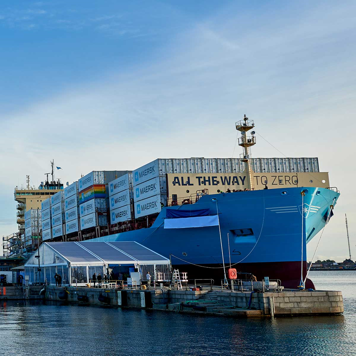Large teal cargo ship, Laura Maersk, docked at a port beside a pier, with containers and a nearby tent structure visible.