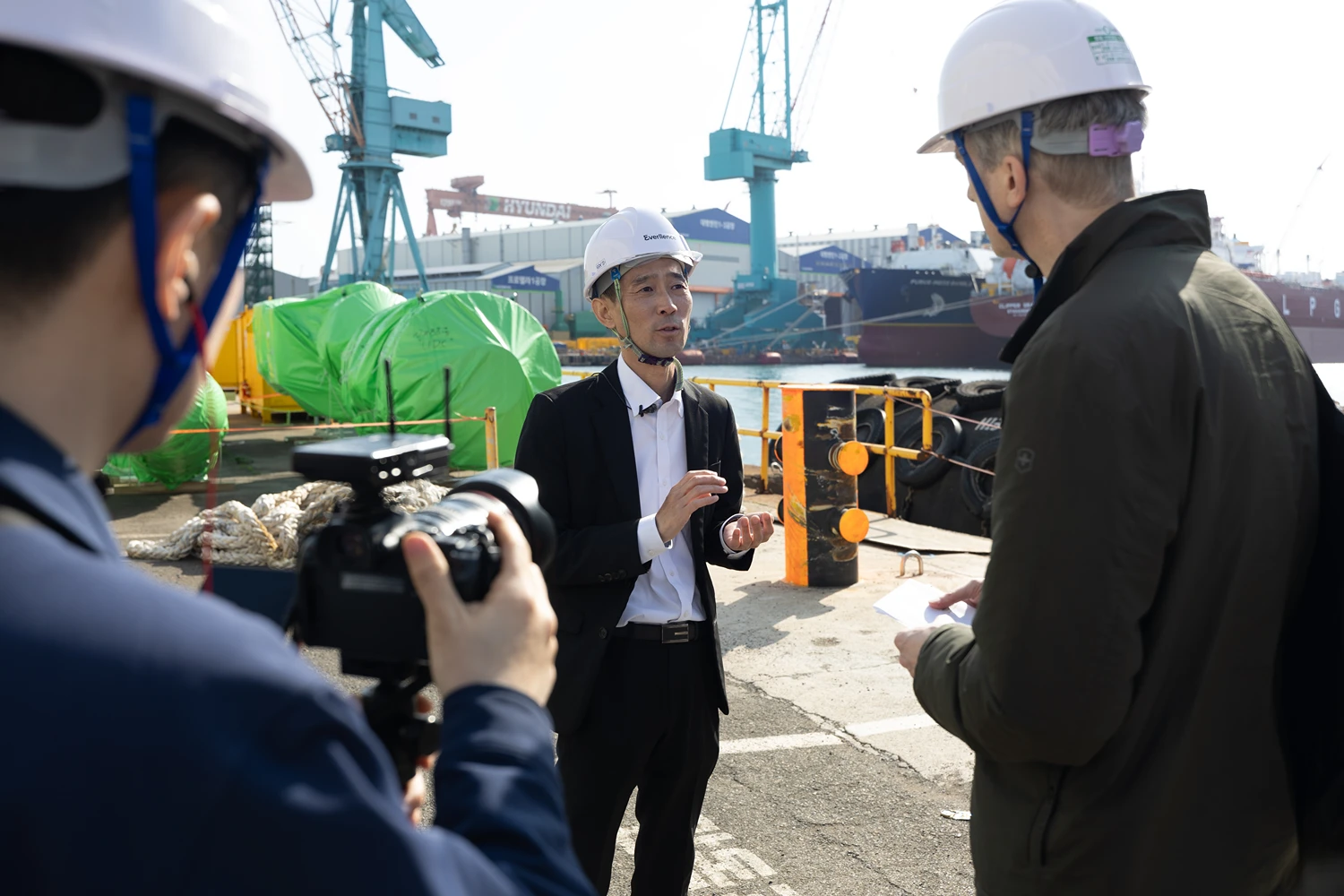 Person wearing safety equipment standing in a shipyard with a large vessel under construction and ammonia engine transport activities visible in the background.