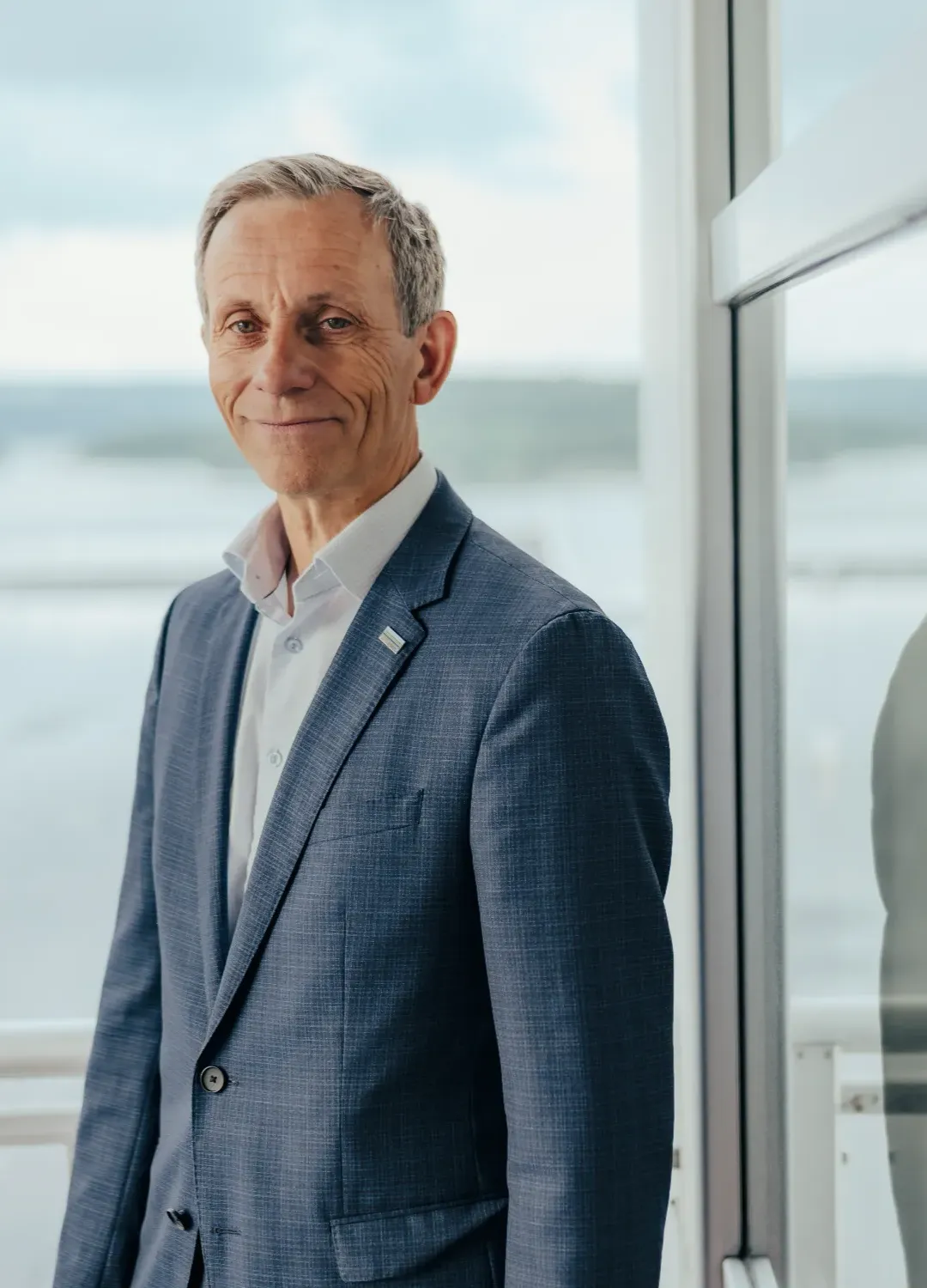 Jon Rysst in a blue suit standing next to a glass window with their face blurred, reflection visible in the glass