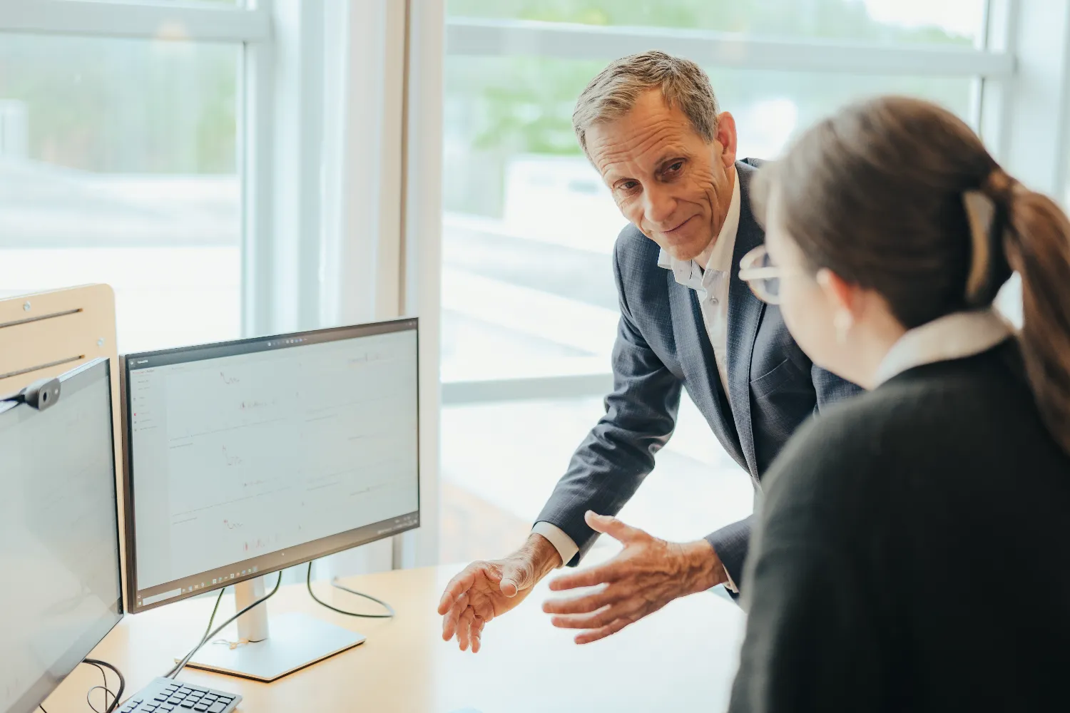 Two professionals in an office setting discussing data displayed on a computer monitor, one standing and gesturing, the other seated