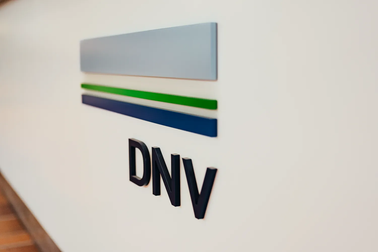 Wall-mounted DNV logo featuring three horizontal bars in light blue, green, and dark blue, with the letters “DNV” below