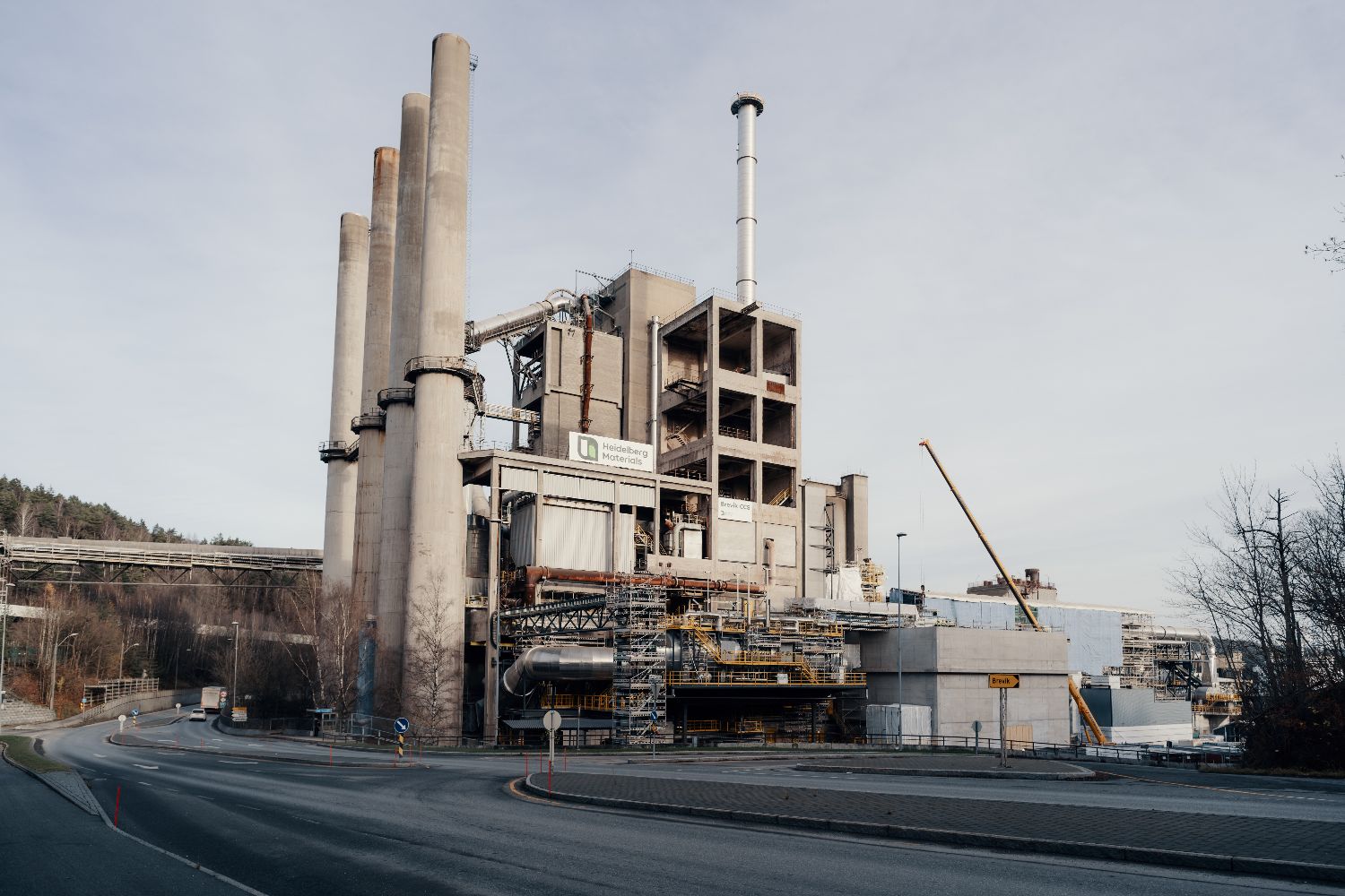 Industrial site featuring large chimneys, scaffolding, and signage for Heidelberg Materials and Brevik CCS, located near a road with trees in the background.