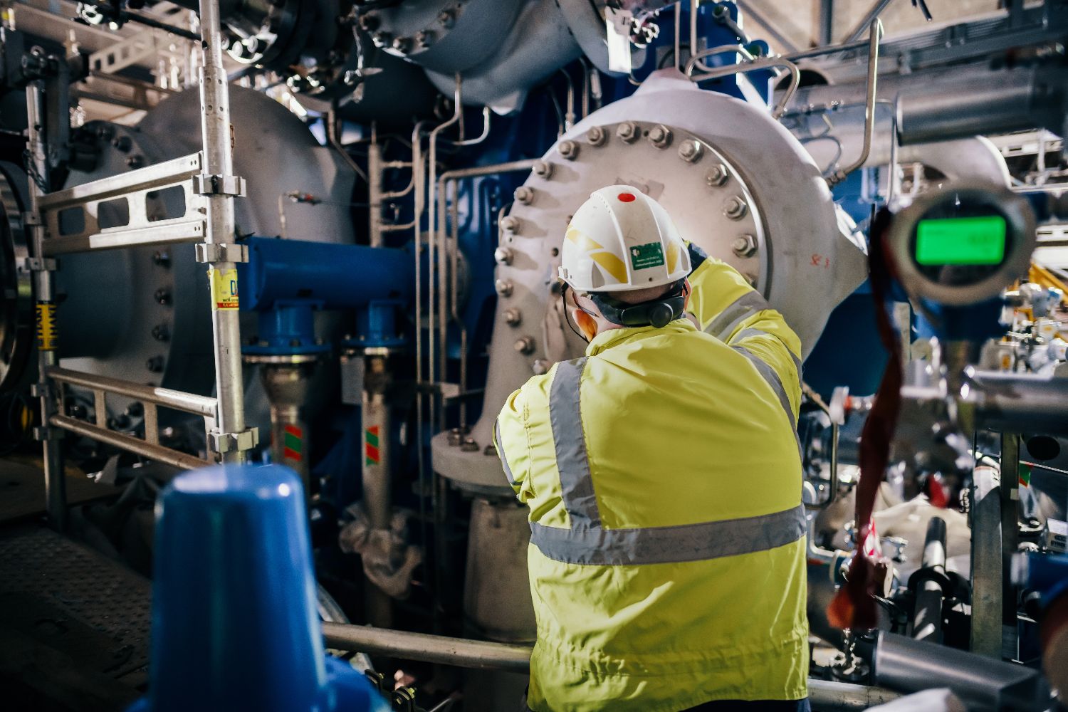 Worker in high-visibility gear and hard hat inspecting industrial carbon capture machinery with pipes and valves at Heidelberg Materials.