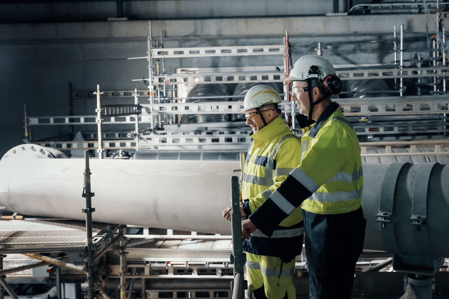 Two workers in high-visibility safety gear and helmets inspecting industrial machinery and pipe systems at a carbon capture site.