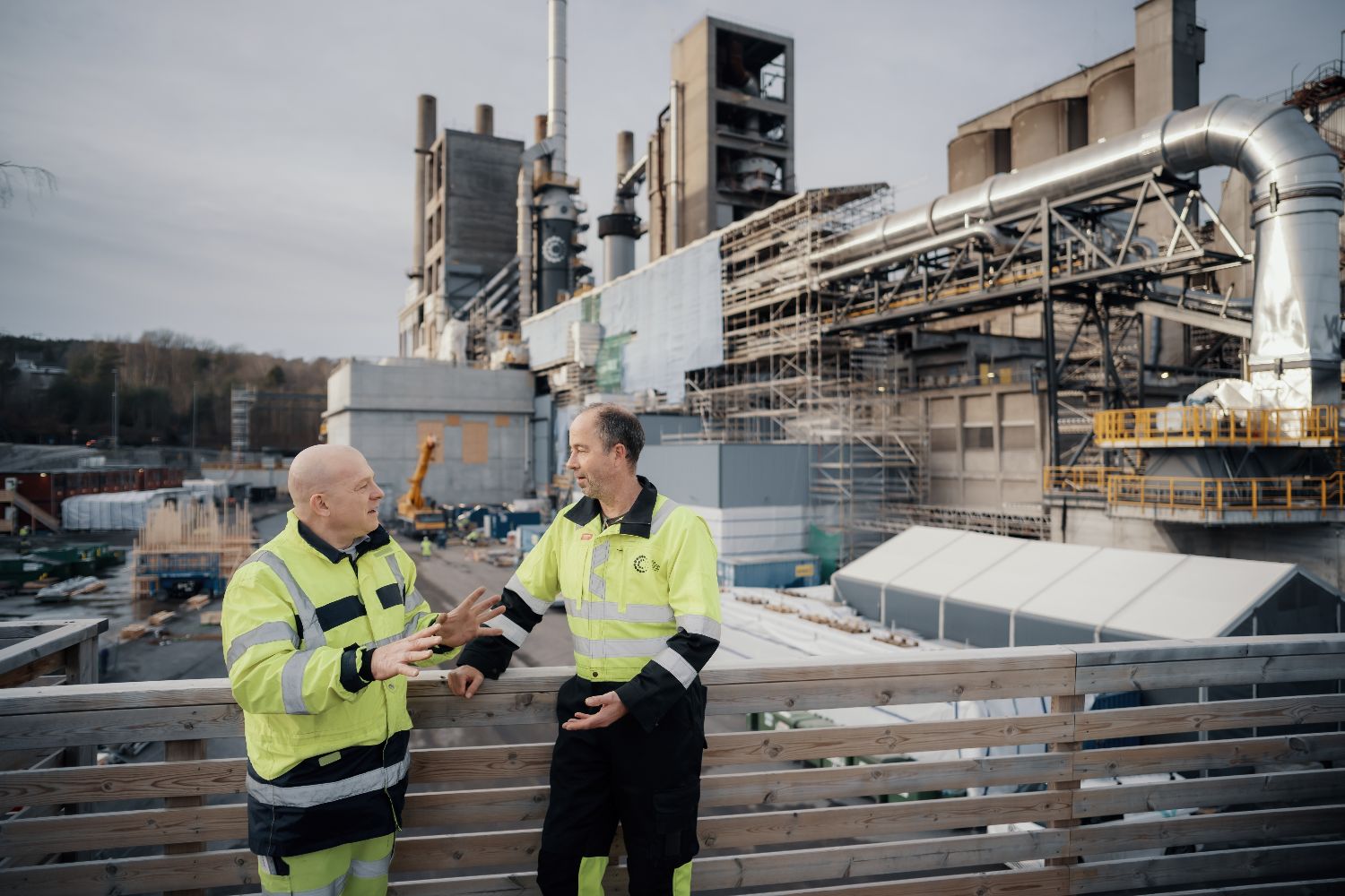 Two individuals in high-visibility jackets standing on a wooden platform, discussing in front of an industrial facility with scaffolding and machinery at a carbon capture site.