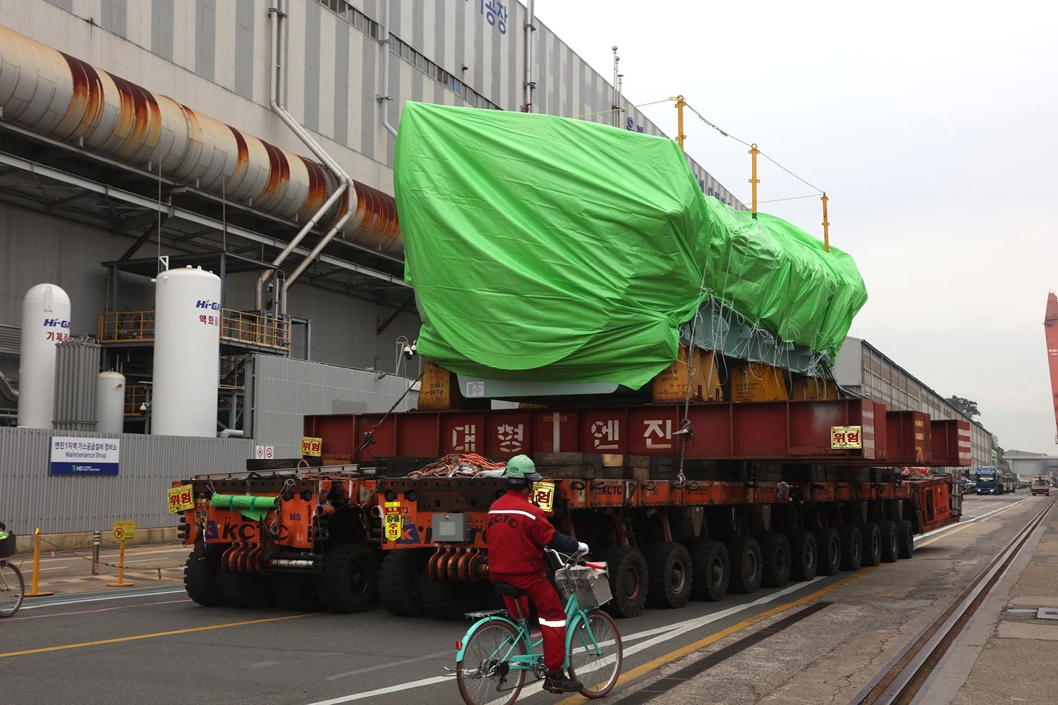 Large engine section wrapped in green protective covering being transported on a multi-axle heavy-load carrier through a shipyard.