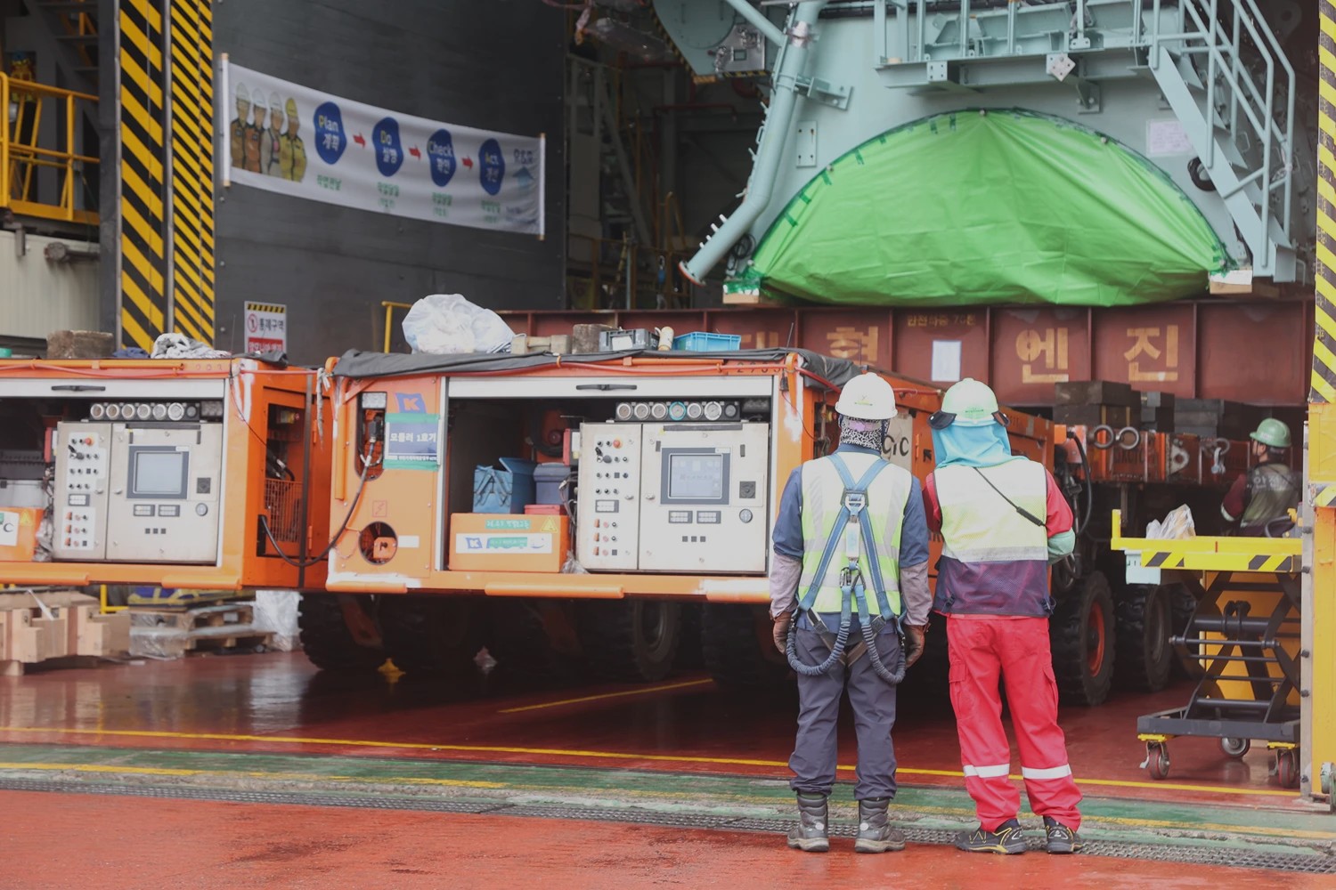 Two workers standing in front of a large ammonia engine module covered with green protective sheeting inside a shipyard assembly hall.