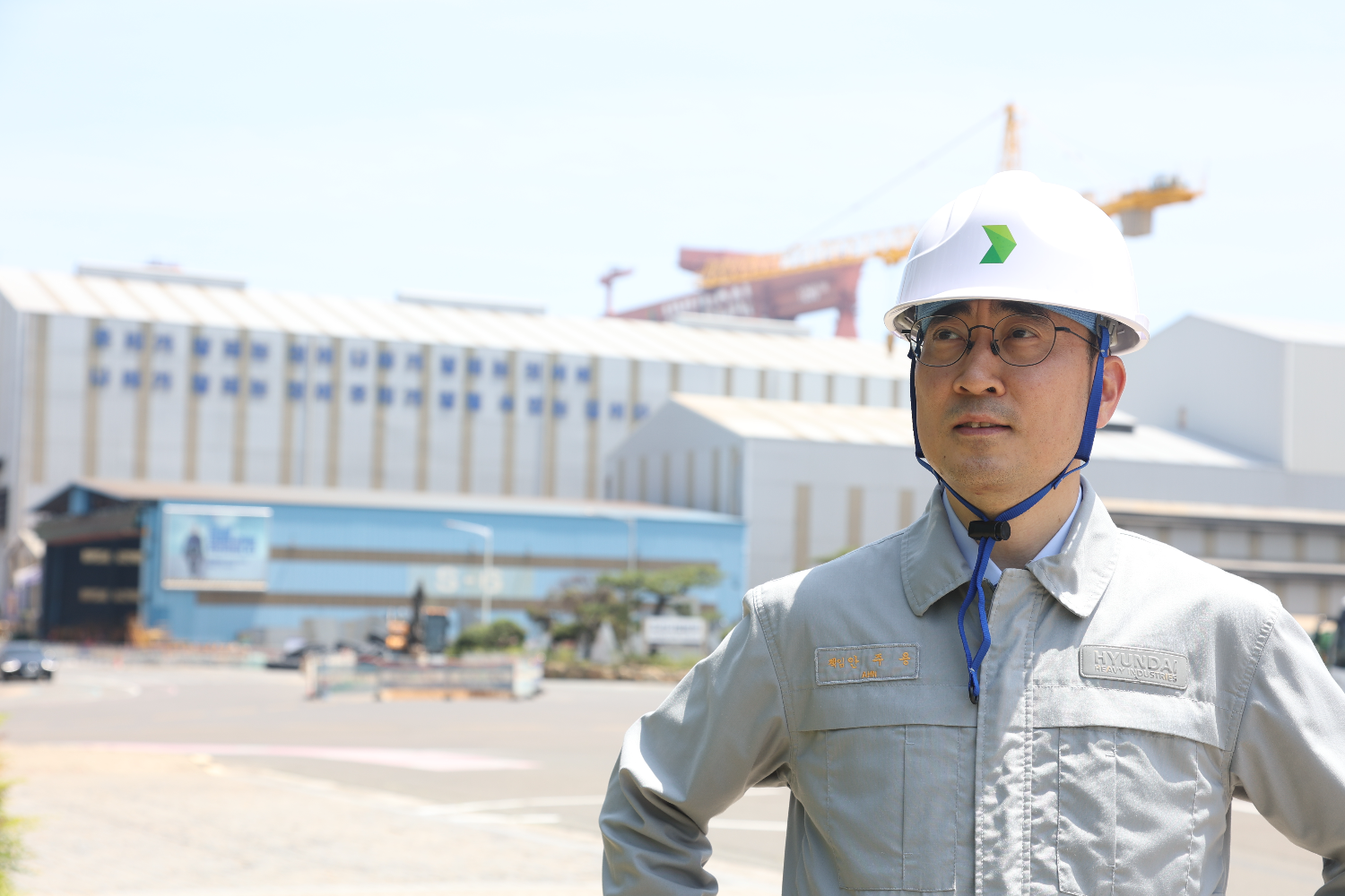 Close-up of a shipyard worker wearing a white safety helmet and grey work uniform with Hyundai branding, standing outdoors in front of large industrial buildings and cranes at Ulsan shipyard in bright daylight