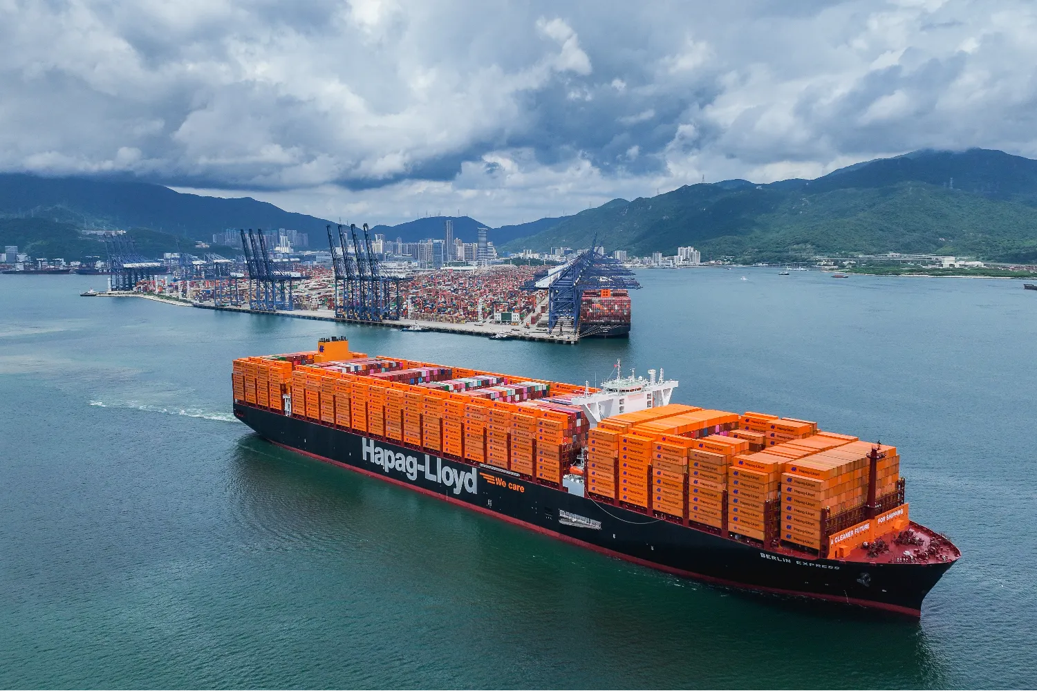 The Berlin Express container ship from Hapag-Lloyd sailing near a port, loaded with orange containers and surrounded by cranes and mountains in the background