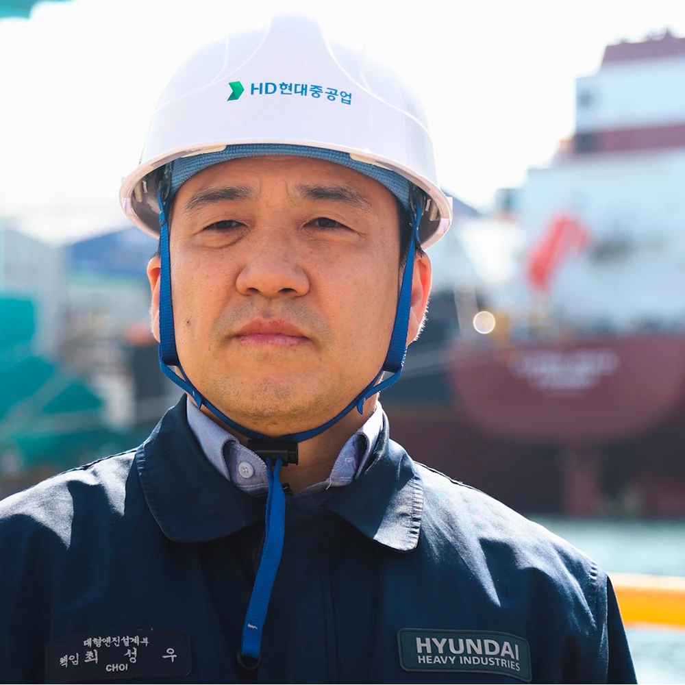 Person wearing a white safety helmet and dark work jacket standing in front of a shipyard with a vessel visible in the background.