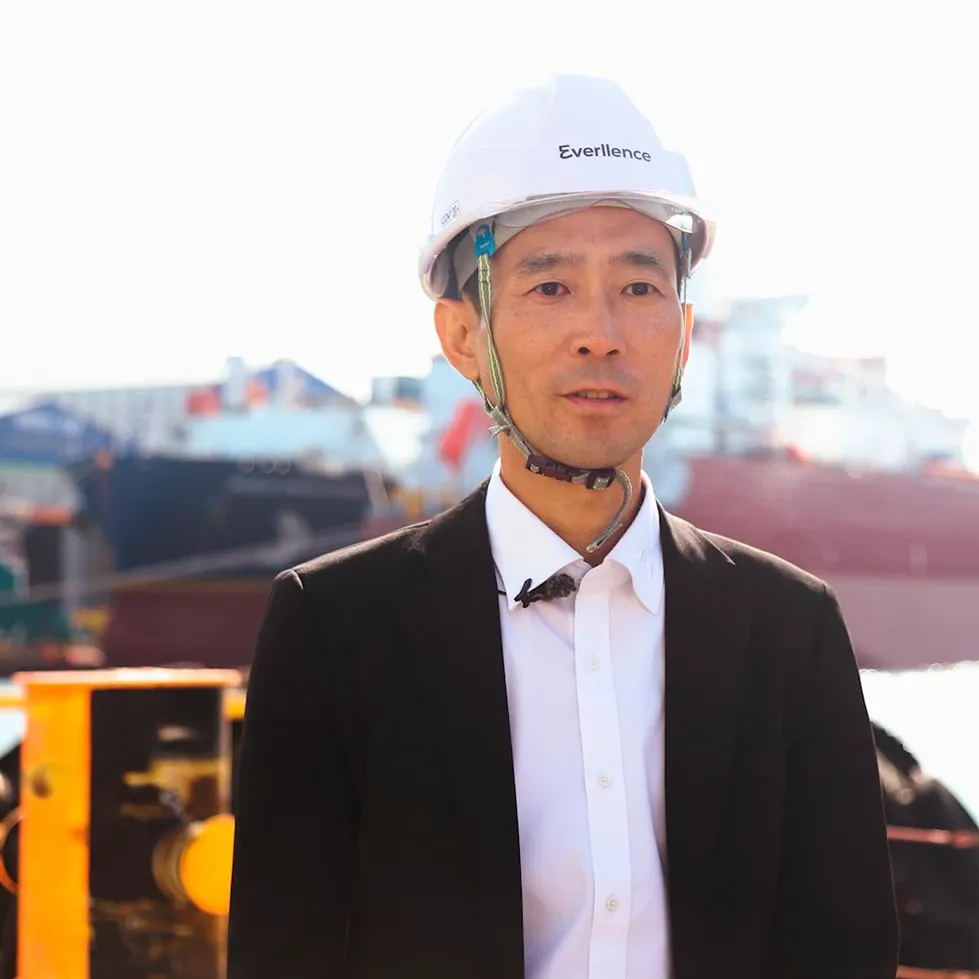 Person wearing a white safety helmet, dark blazer, and safety vest standing at a shipyard with industrial structures and vessels behind.