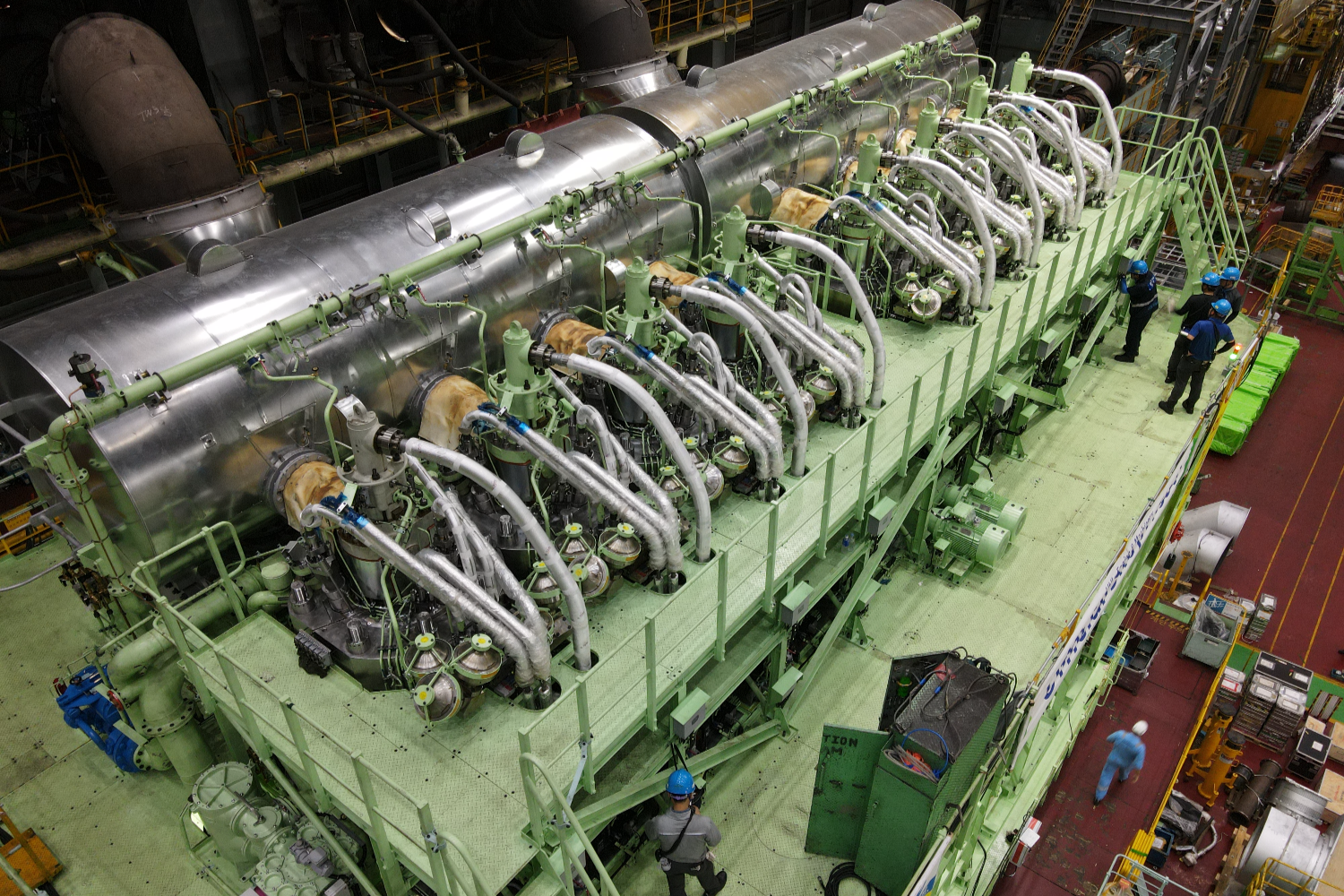 Top-down view of a long two-stroke marine engine with multiple cylinders aligned in a row, surrounded by walkways and industrial equipment