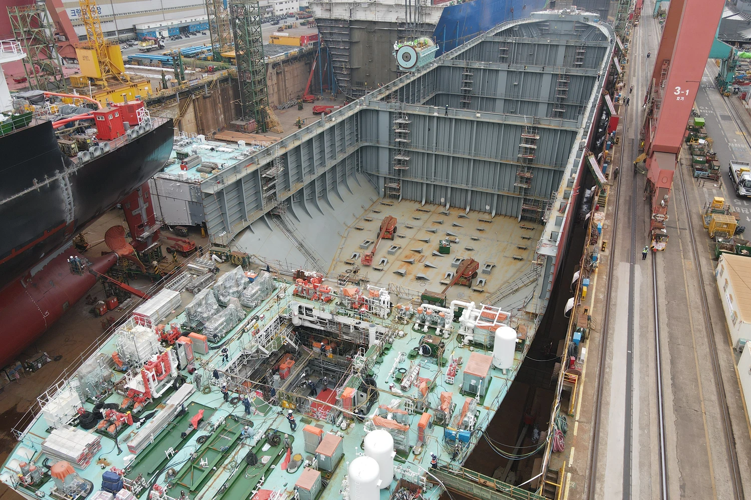 Aerial view into a large vessel hull where engine components are being installed in the engine room beneath towering ship structures.