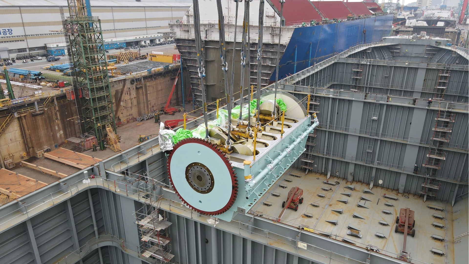 Large ammonia engine section suspended by crane and being lowered into the open hull of a vessel under construction at a shipyard, with workers and heavy lifting equipment visible below.