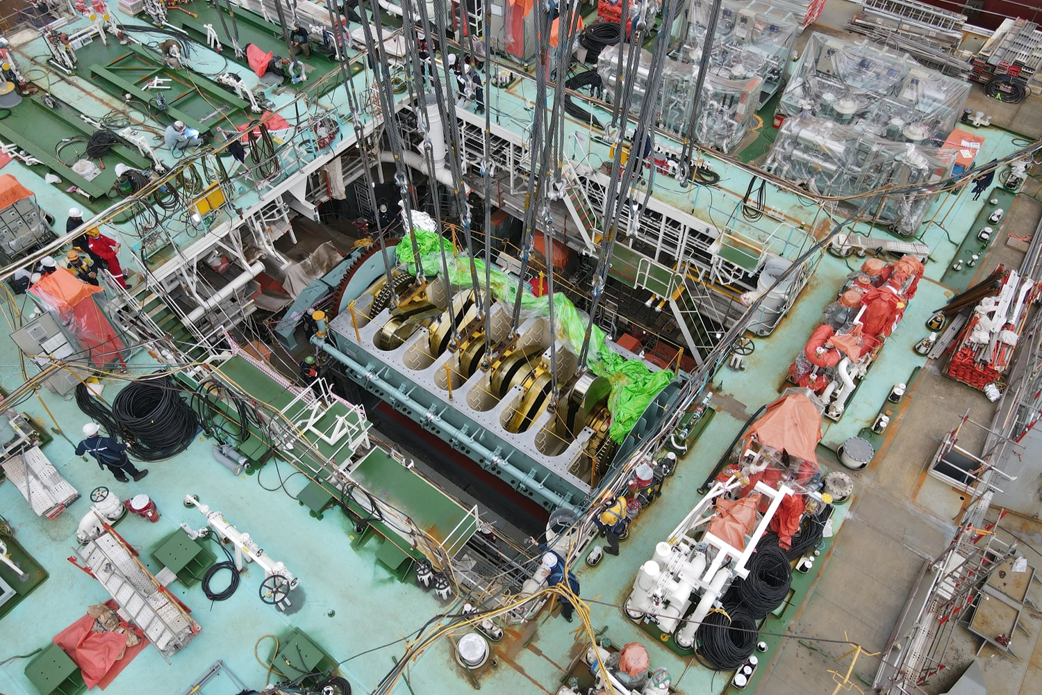 Heavy engine component suspended by crane above a vessel engine room during installation inside a partially completed ship hull.