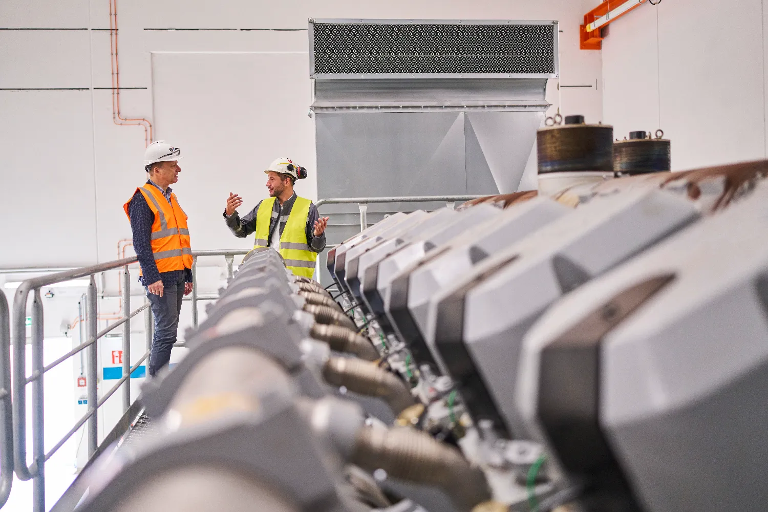 Two project managers wearing safety helmets and high-visibility vests stand on a platform inside the Chemnitz-Nord CHP plant, discussing operations in front of large industrial machinery and piping.