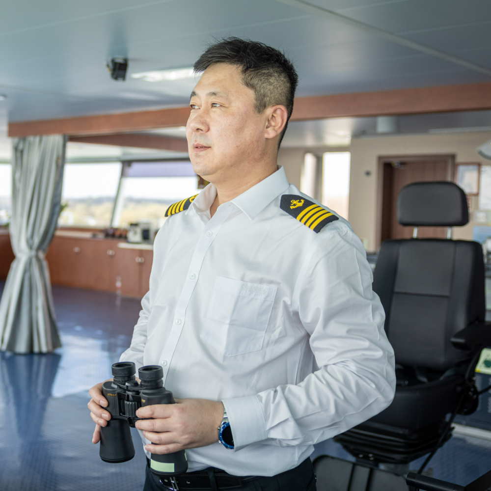 Person standing on a ship bridge in front of large windows, with control panels and navigation equipment visible, overlooking the sea during daylight