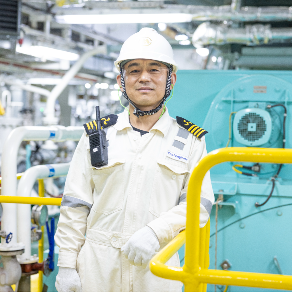 Person standing inside an engine room next to large turquoise-colored machinery and pipes, wearing protective clothing and safety helmet