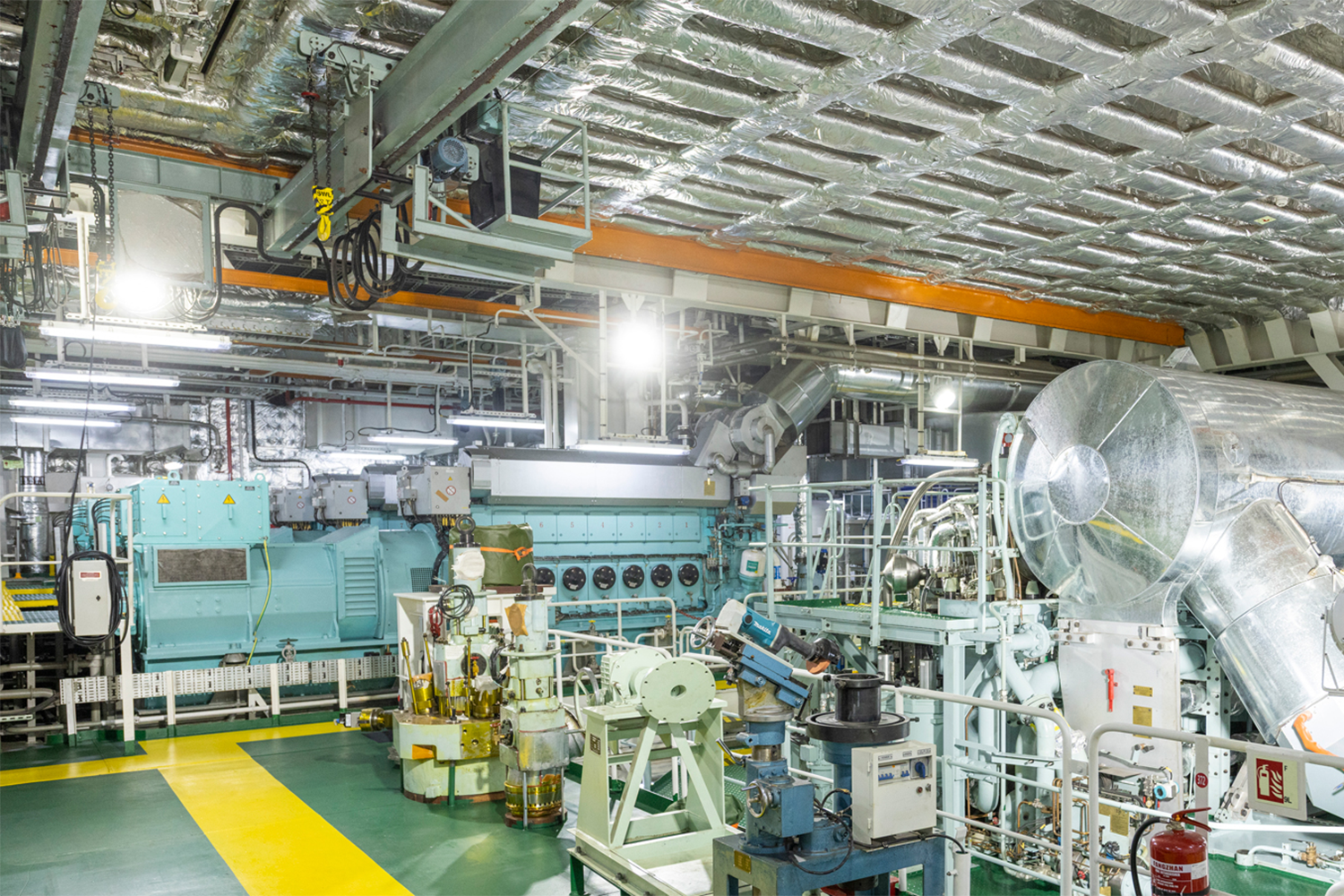 Interior of a ship engine room with a large turquoise main engine, surrounded by walkways, pipes, and overhead structures