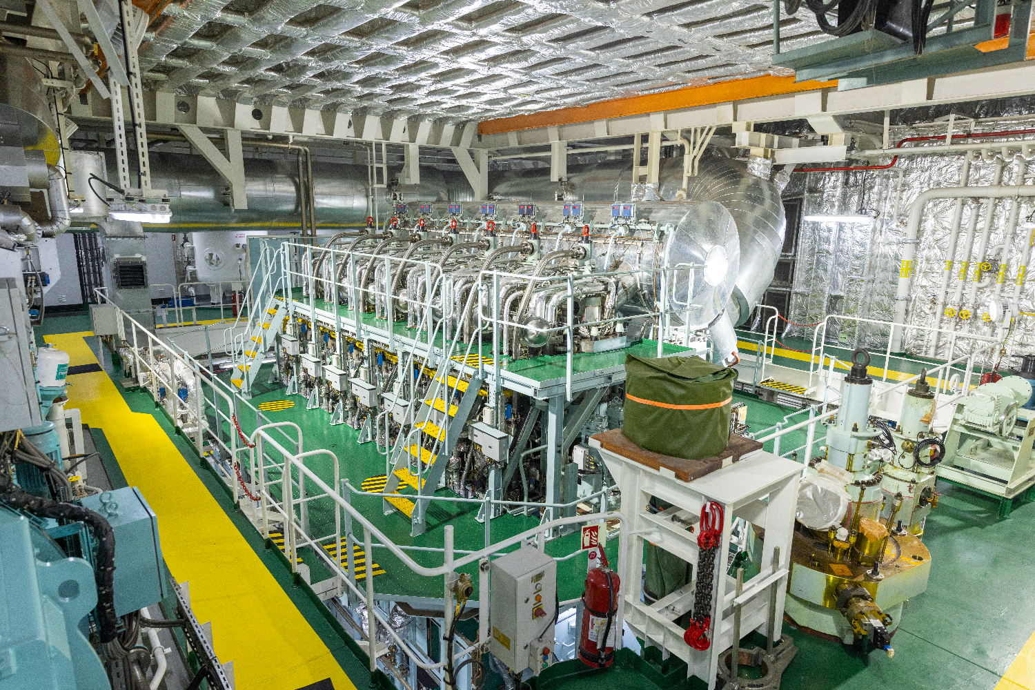 Engine room view showing multiple smaller generator units arranged in a row, with piping, platforms, and industrial lighting overhead