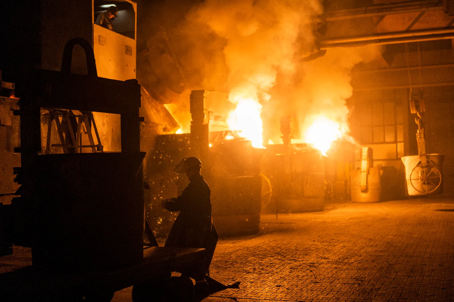 A foundry worker in protective gear stands near glowing furnaces as molten iron is prepared for casting in the Everllence foundry in Augsburg, Germany.