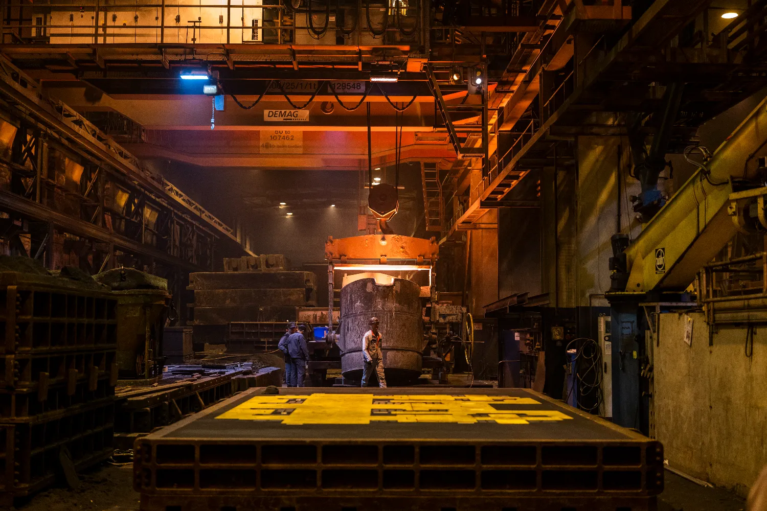 A large crucible filled with molten iron is suspended from a DEMAG overhead crane, with foundry workers in protective gear overseeing the process inside the Everllence foundry in Augsburg.