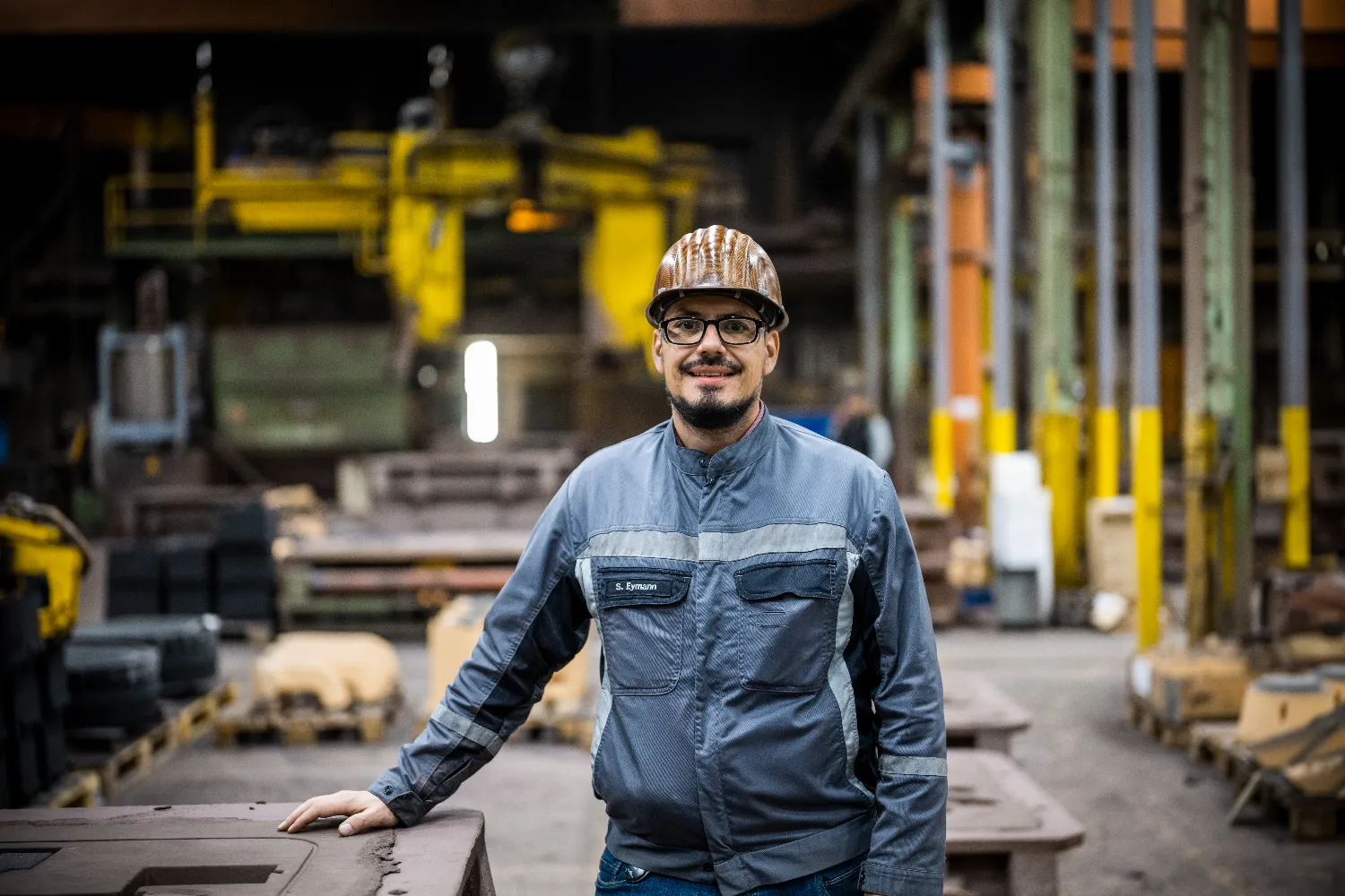 Sven Eymann, wearing a hard hat and safety gear, stands inside the Everllence foundry in Augsburg, surrounded by industrial equipment and structural elements.