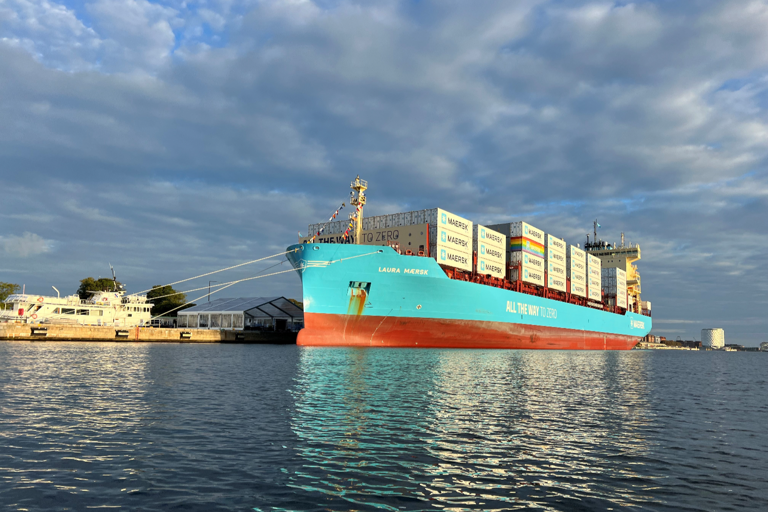 Large container vessel sailing on open water with stacked containers on deck and a coastal city visible in the background