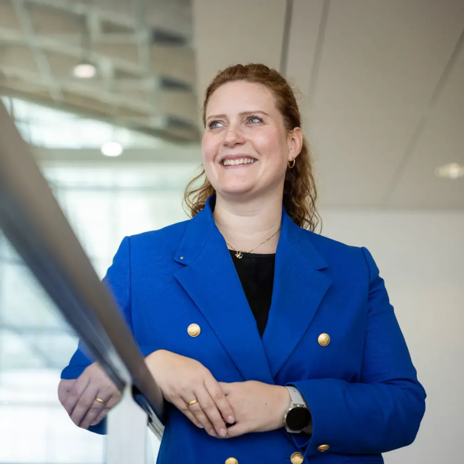 Silke Lehmköster in a blue blazer with gold buttons standing indoors in front of large windows and modern architectural elements, face blurred