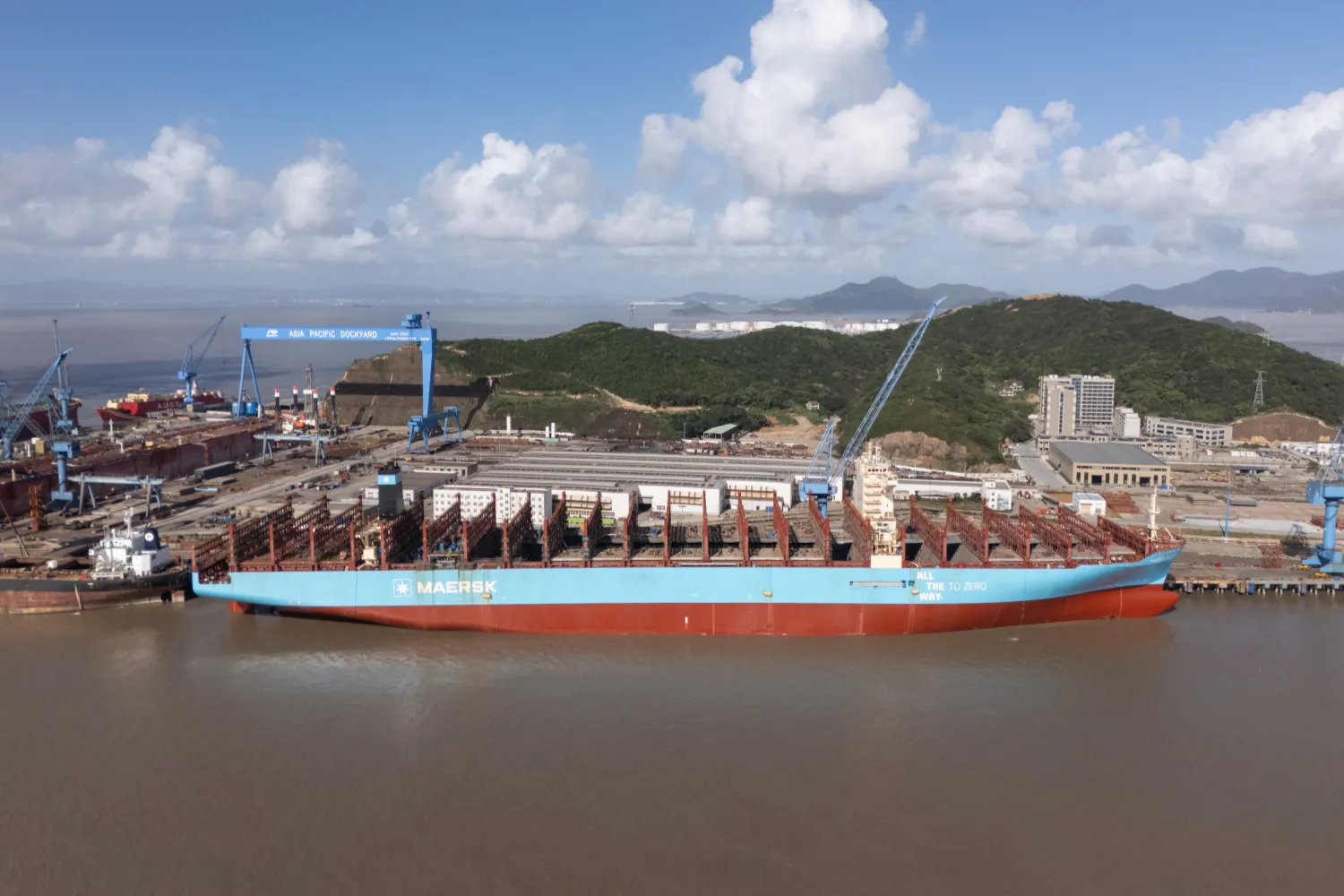 Container ship with “ALL THE WAY TO ZERO” slogan docked at Asia Pacific Dockyard, surrounded by cranes, equipment, and green hills in the background.