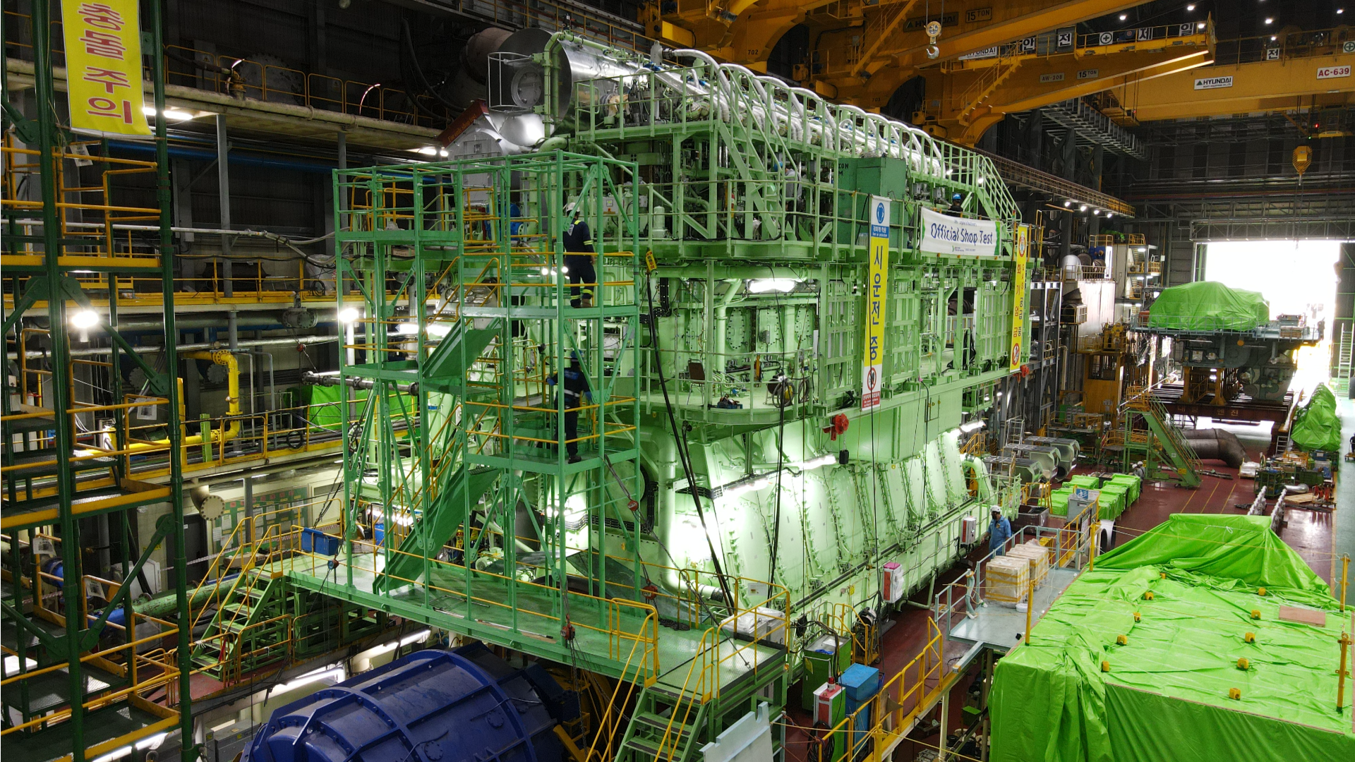 Large green-painted marine engine inside a production hall, surrounded by cranes, platforms, and industrial lighting during assembly