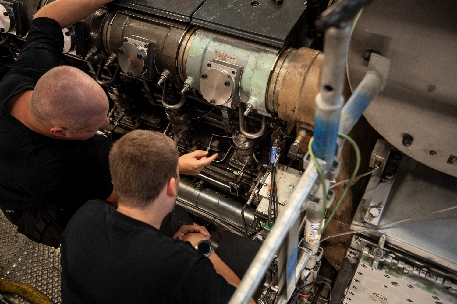 Two engineers leaning over a marine engine, closely examining piping, injectors, and internal components in a compact engine space
