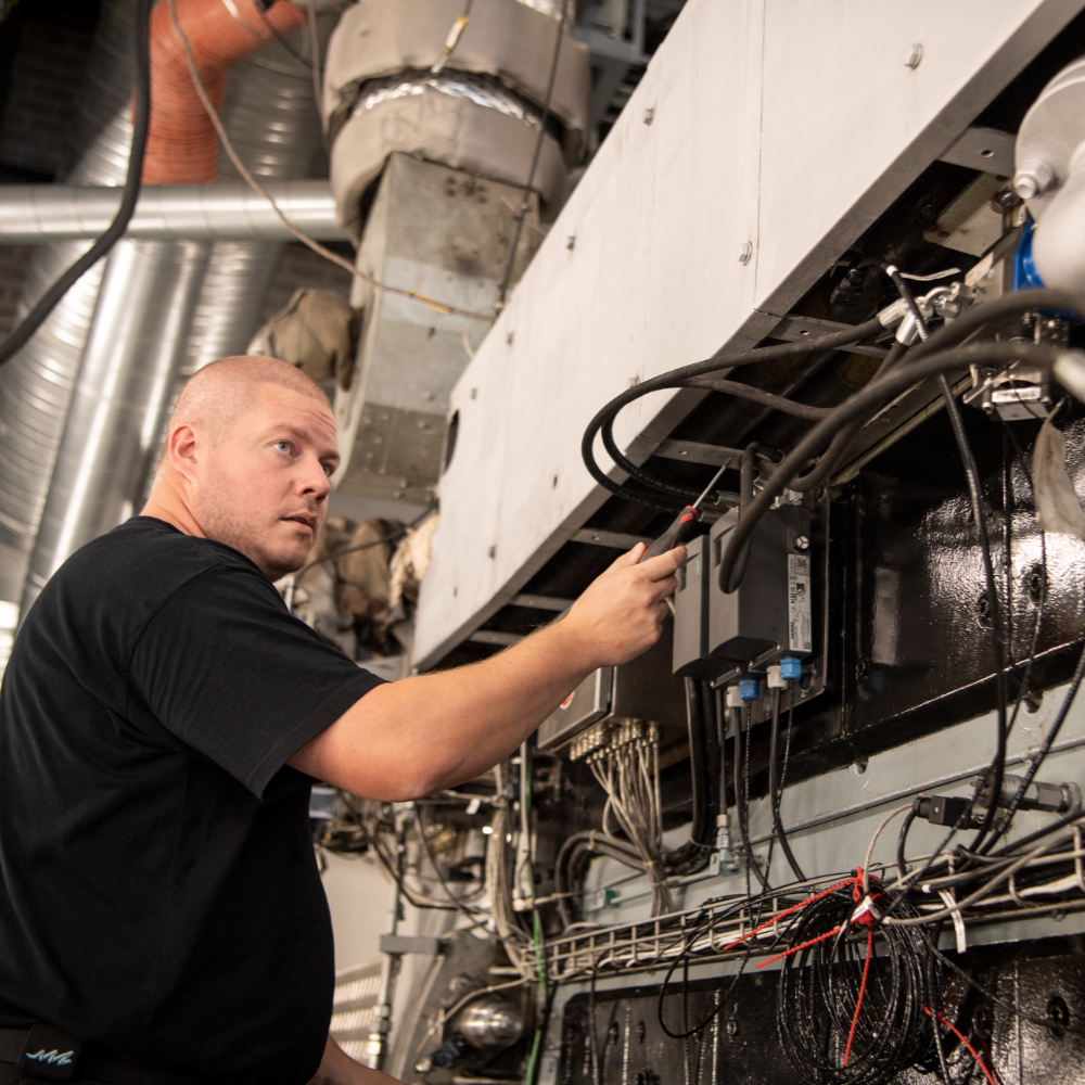 Engineer standing and working on the side of a large engine, interacting with cables, pipes, and mounted equipment in a test hall