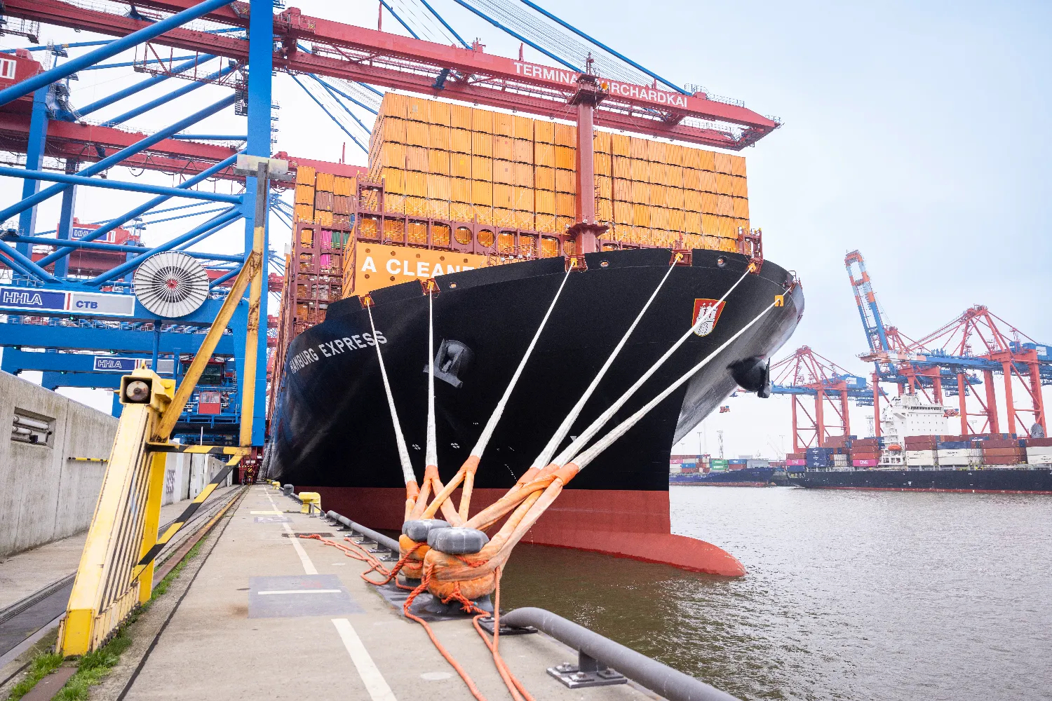 The Hamburg Express docked at Terminal Burchardkai in Hamburg, secured with mooring lines and surrounded by large container cranes.