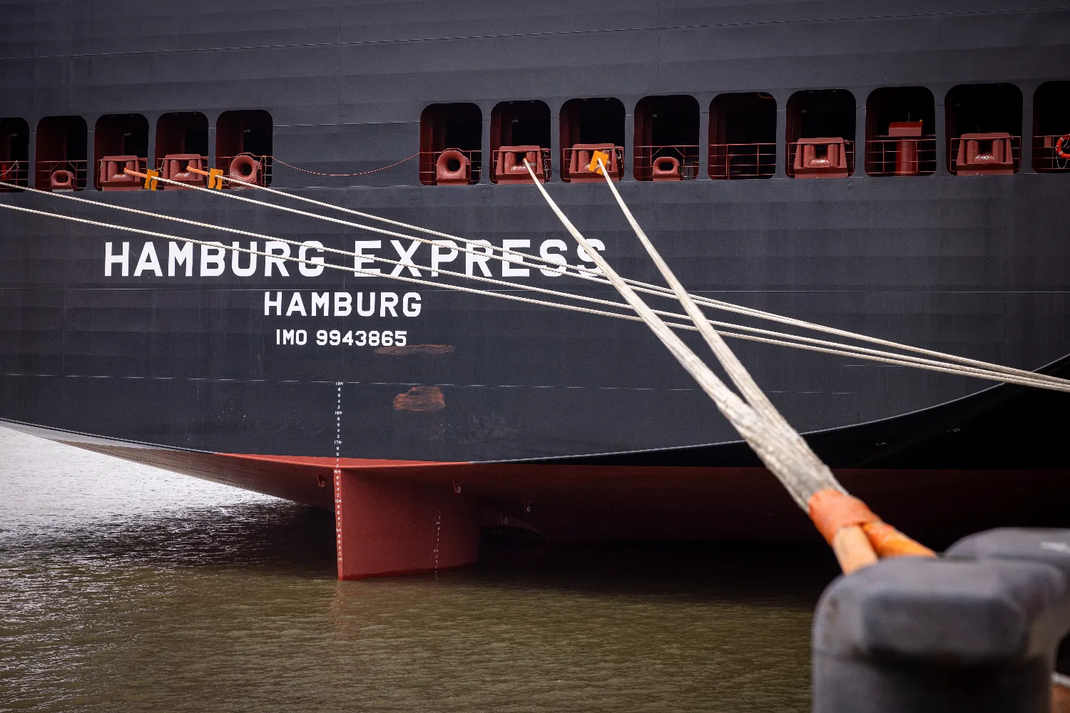 Close-up of the Hamburg Express’s hull showing its name, home port Hamburg, and IMO number 9943865, with mooring lines securing the vessel at dock.