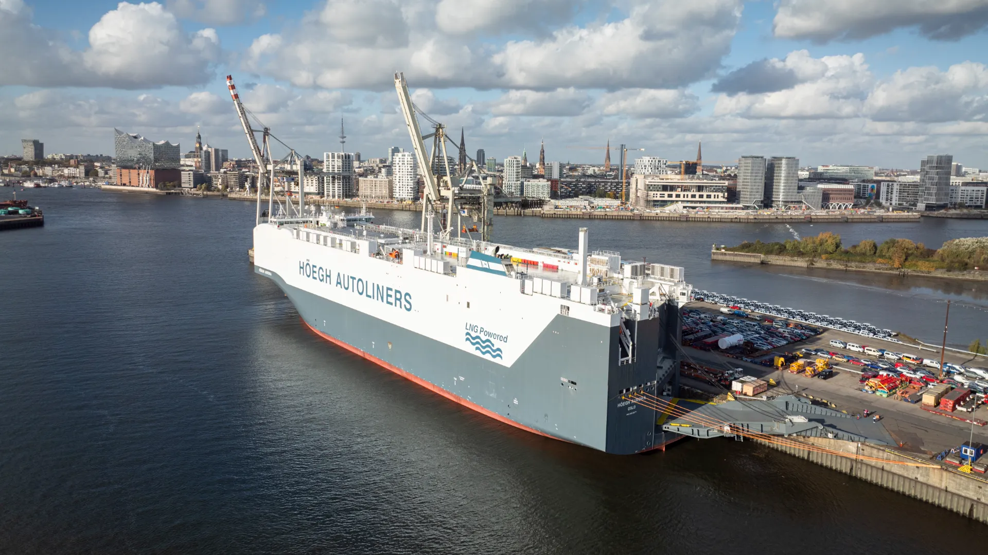 The Höegh Aurora, the world’s largest LNG-powered car carrier, docked at the Port of Hamburg with city buildings in the background and cargo vehicles nearby.