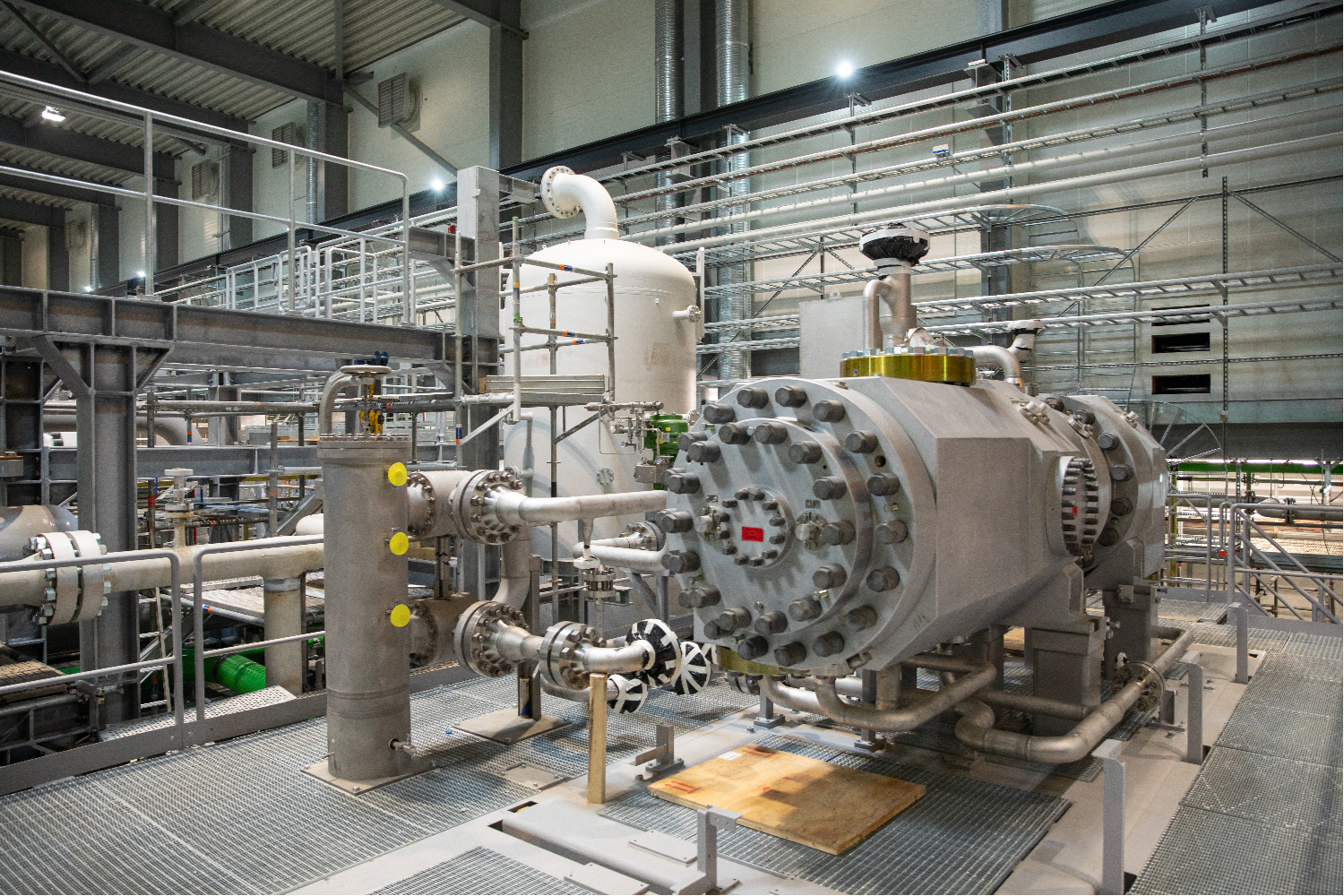 Large industrial heat pump unit installed inside a high-ceiling hall, surrounded by metal platforms, pipes, and access ladders, with structured piping systems and machinery components on multiple levels