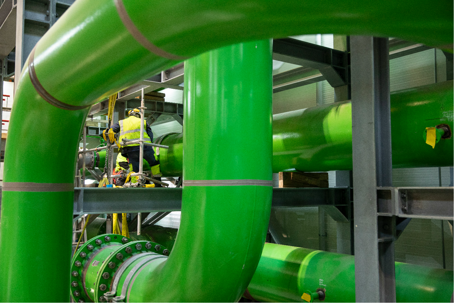 Close-up of bright green industrial pipes running vertically and horizontally through a plant structure, showing valves, connectors, and dense piping layout inside a technical facility