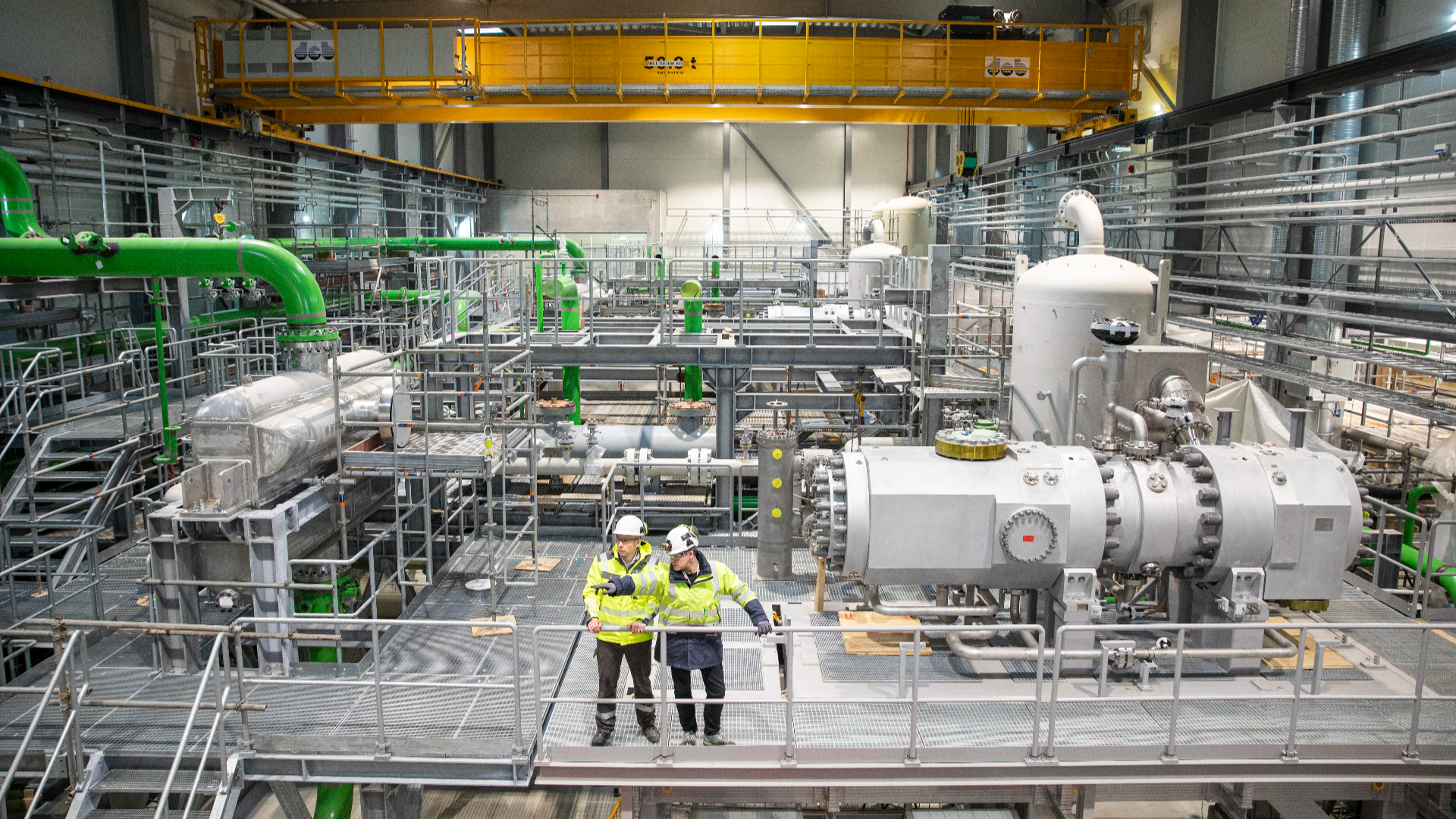 Wide view of a large industrial hall with multiple heat pump units, piping systems, and two workers in safety gear standing on the floor beneath overhead cranes and steel structures