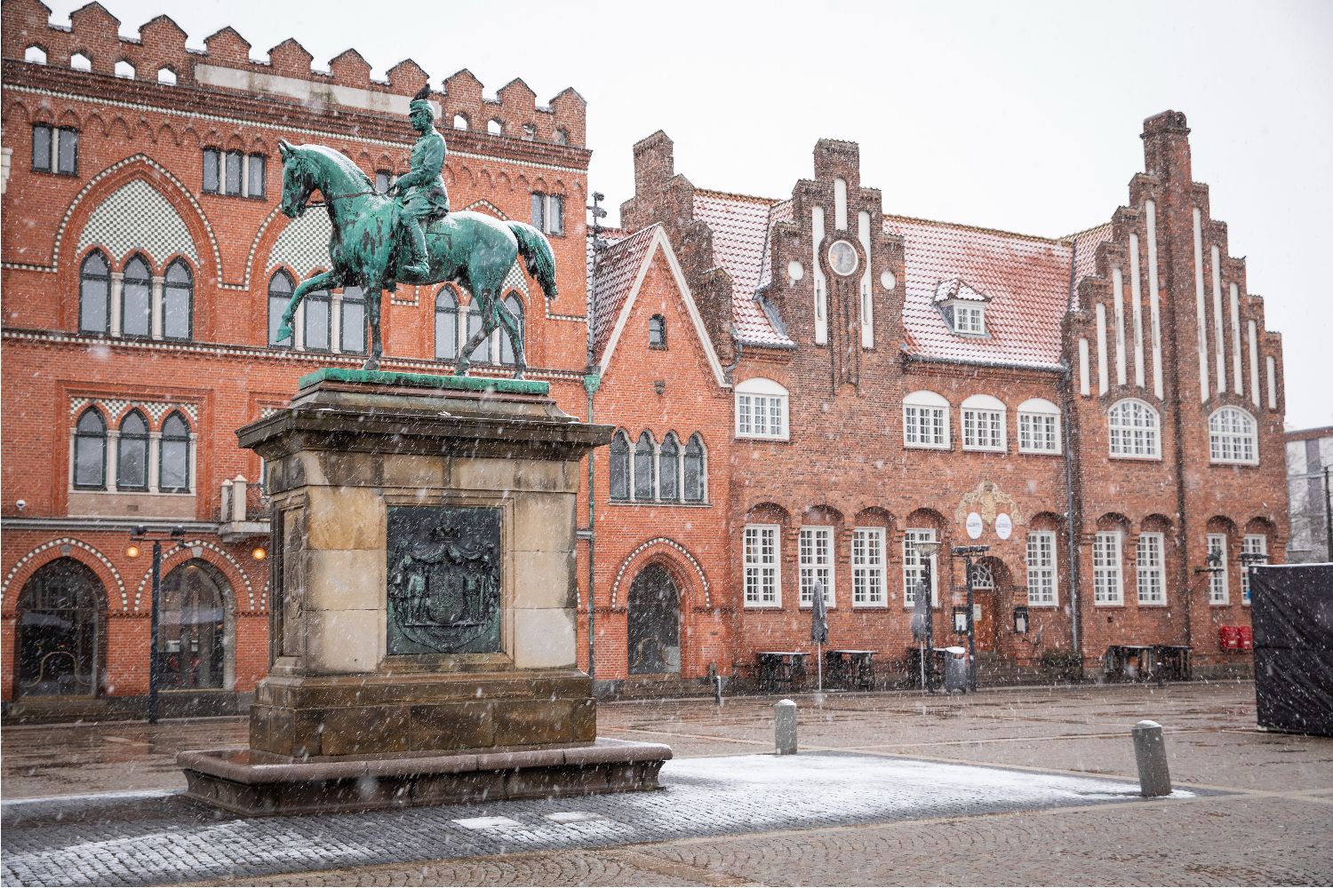 Historic brick building with arched windows and decorative façade in an urban square, with a statue in the foreground and paved open space