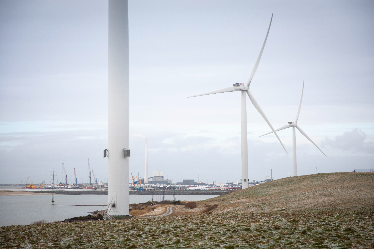 Offshore wind turbines standing near a coastline under overcast sky, with calm water and distant industrial structures visible along the shore