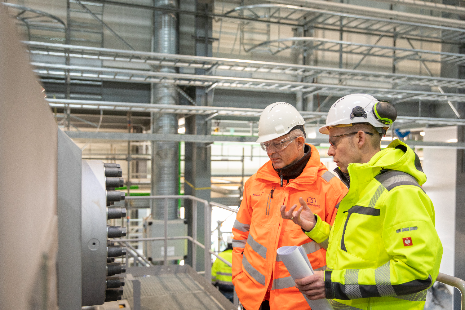 Two workers wearing high-visibility jackets and safety helmets standing next to large industrial pipes and equipment, inspecting components inside a facility