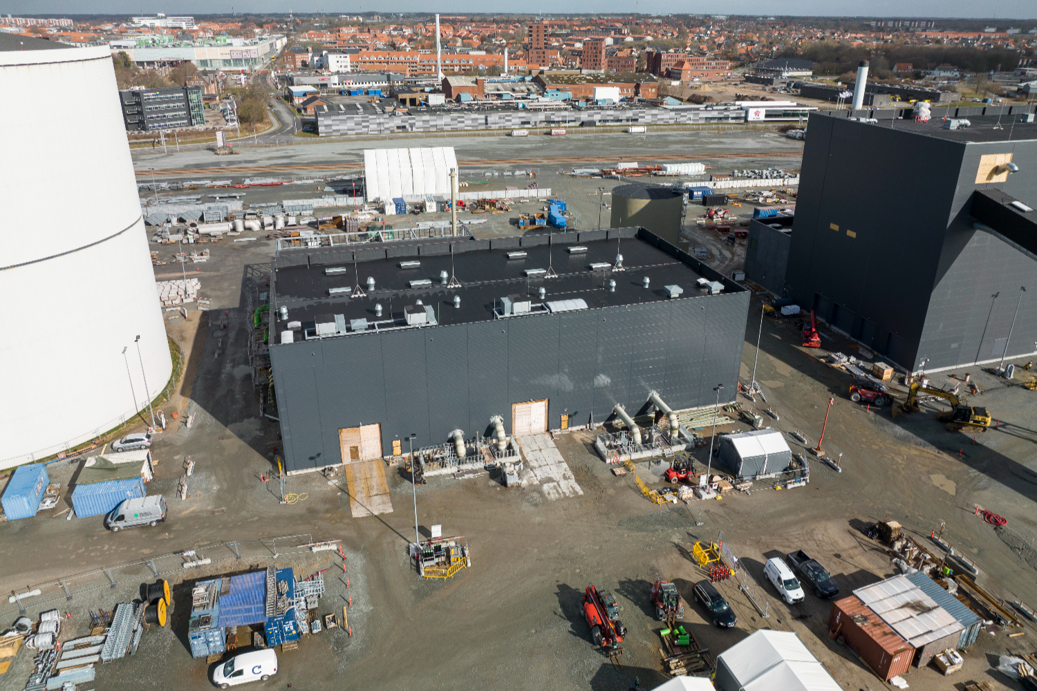 Aerial view of an industrial site with large dark buildings, storage areas, and surrounding infrastructure, located near a harbor and urban coastline