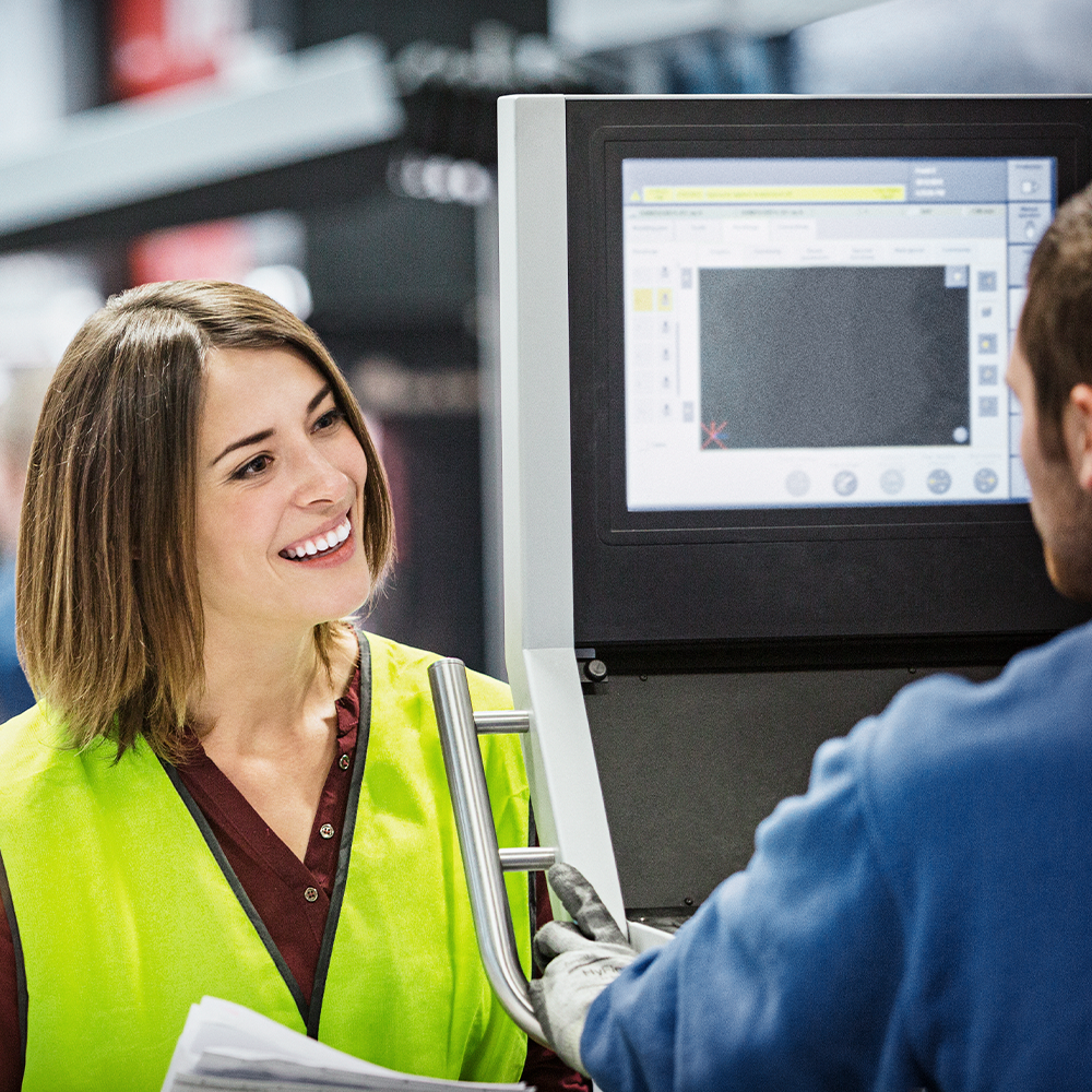 Woman in safety vest smiling while talking to colleague at machine