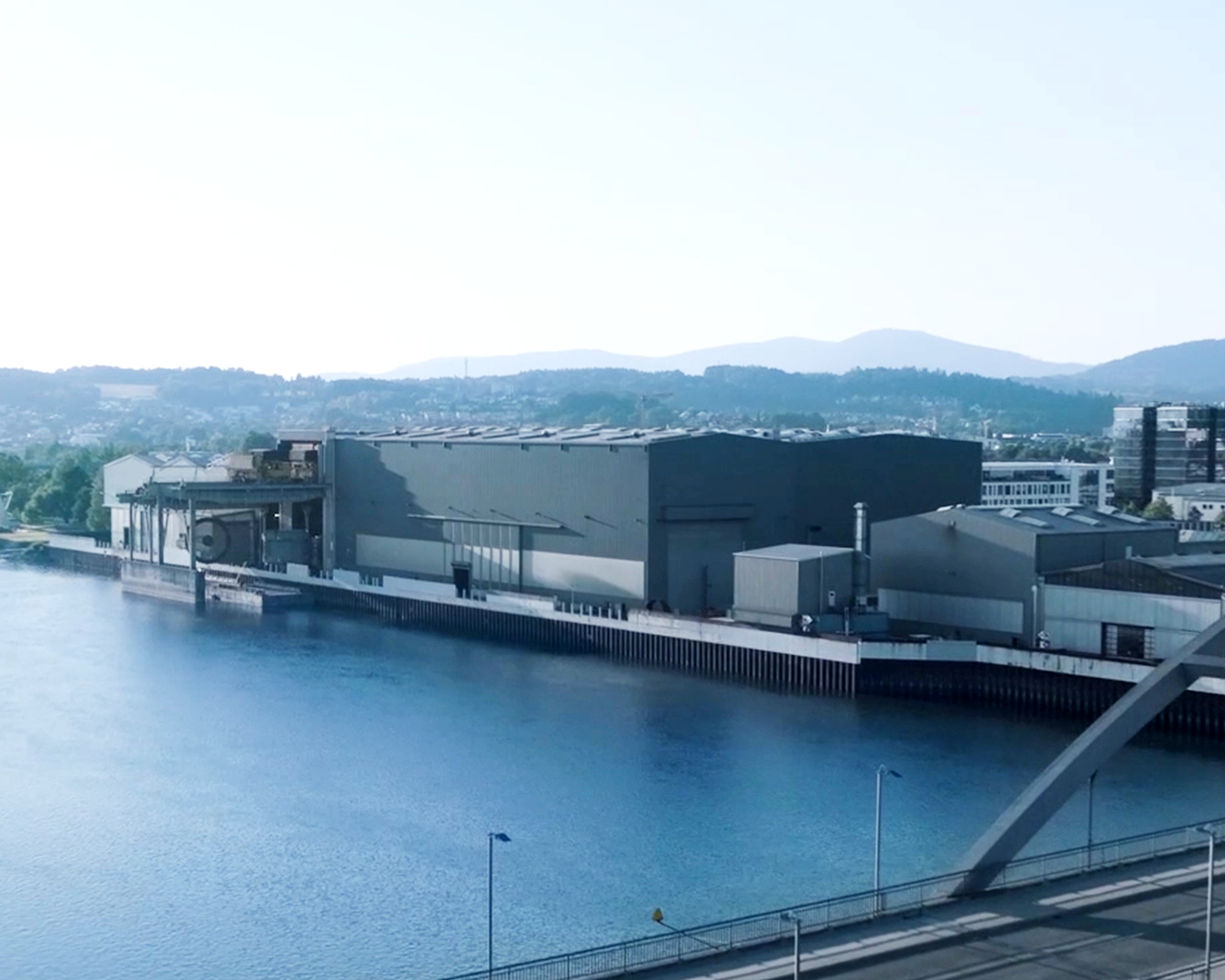 Large industrial hall on a riverbank with bridge and mountains in the background