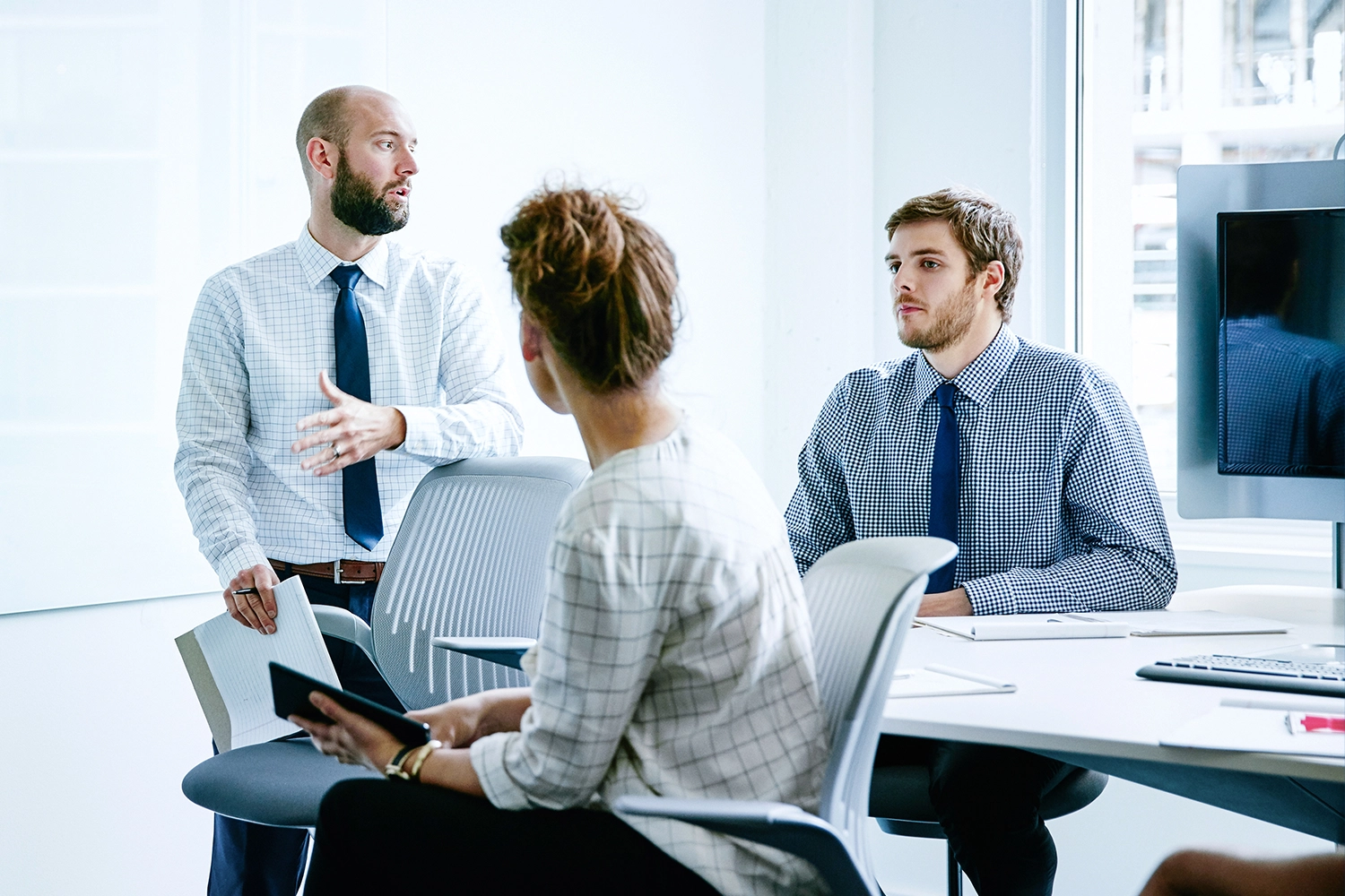 Three people in business attire sitting in a bright office, one man standing and gesturing while others listen