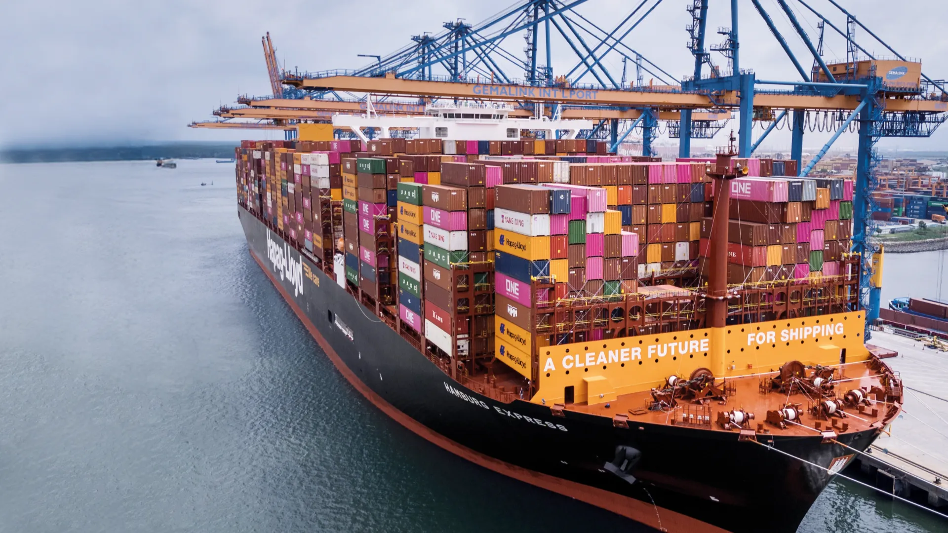 The Hamburg Express container ship docked at Gemalink International Port, loaded with colorful containers and bearing the slogan “A Cleaner Future for Shipping.”