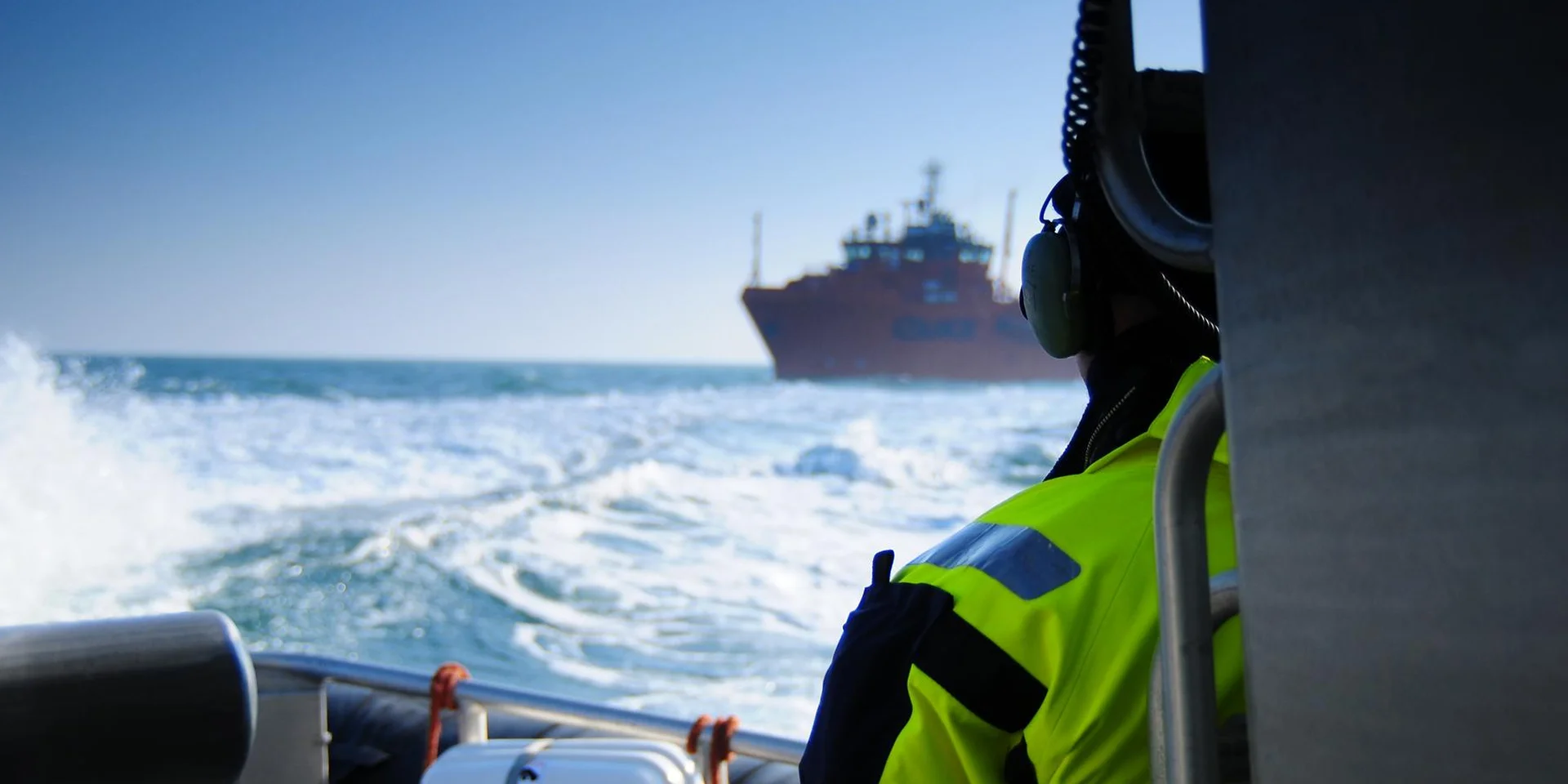 Person wearing a high-visibility jacket on a boat, looking toward a ship at sea